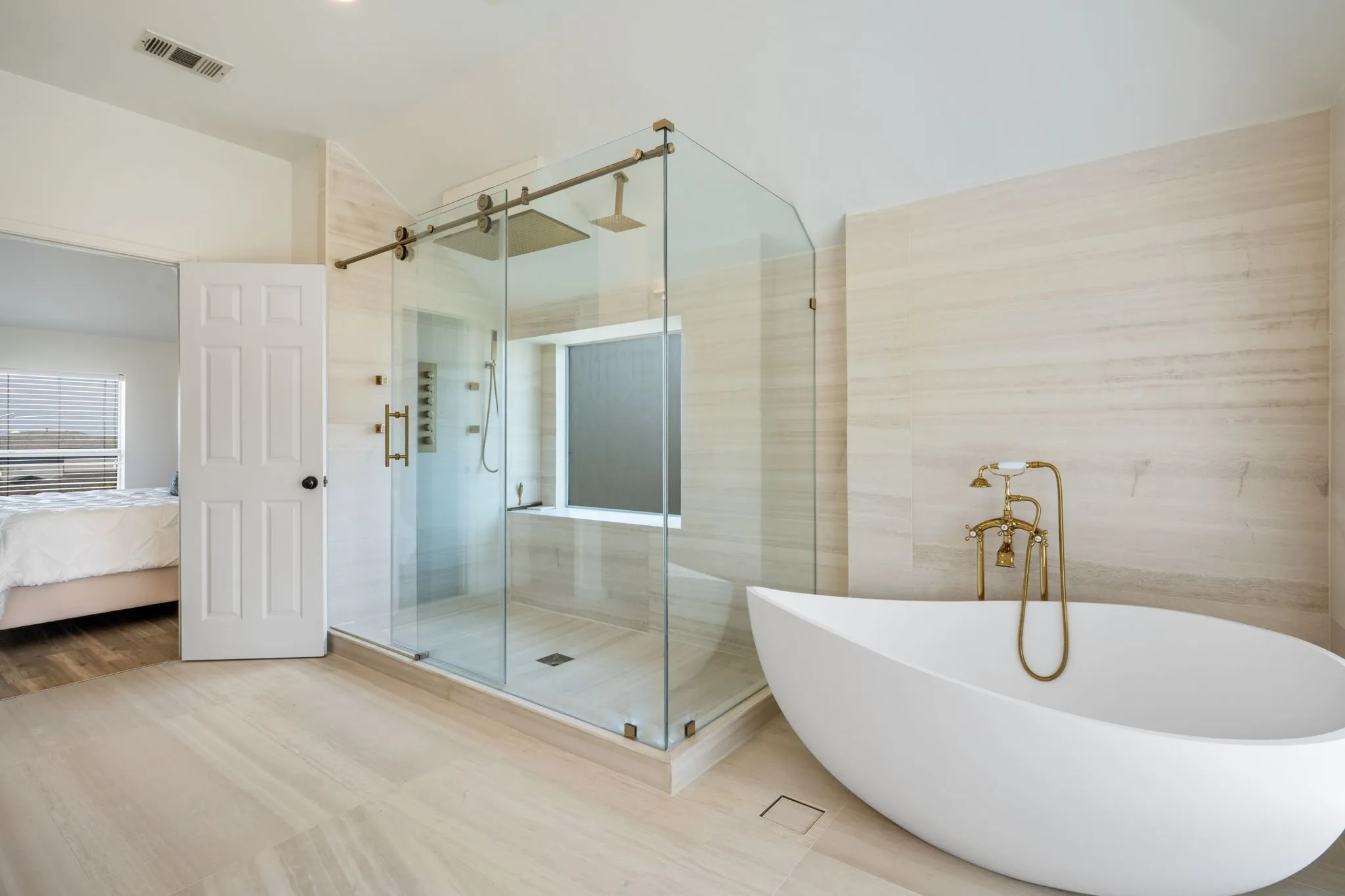 Ensuite presents natural Italian limestone on every wall & floor, a freestanding Japanese soaking tub before a picture window, frameless glass rain shower w body jets, a floating dual vanity beneath a full width LED mirror & a Japanese smart bidet w gold fixtures throughout.