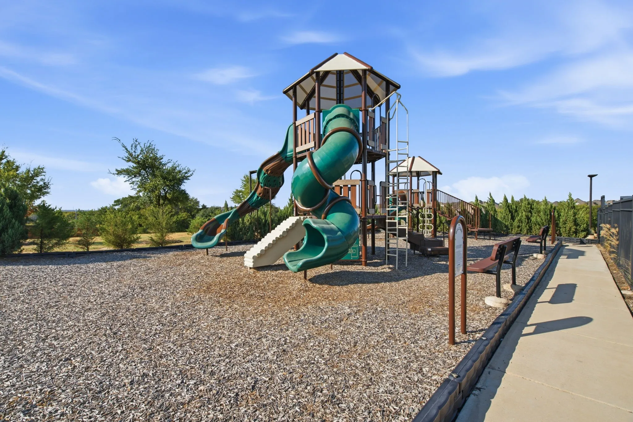 Playground area featuring a multi-level structure with slides and climbing elements, wood chip surfacing, and a concrete pathway