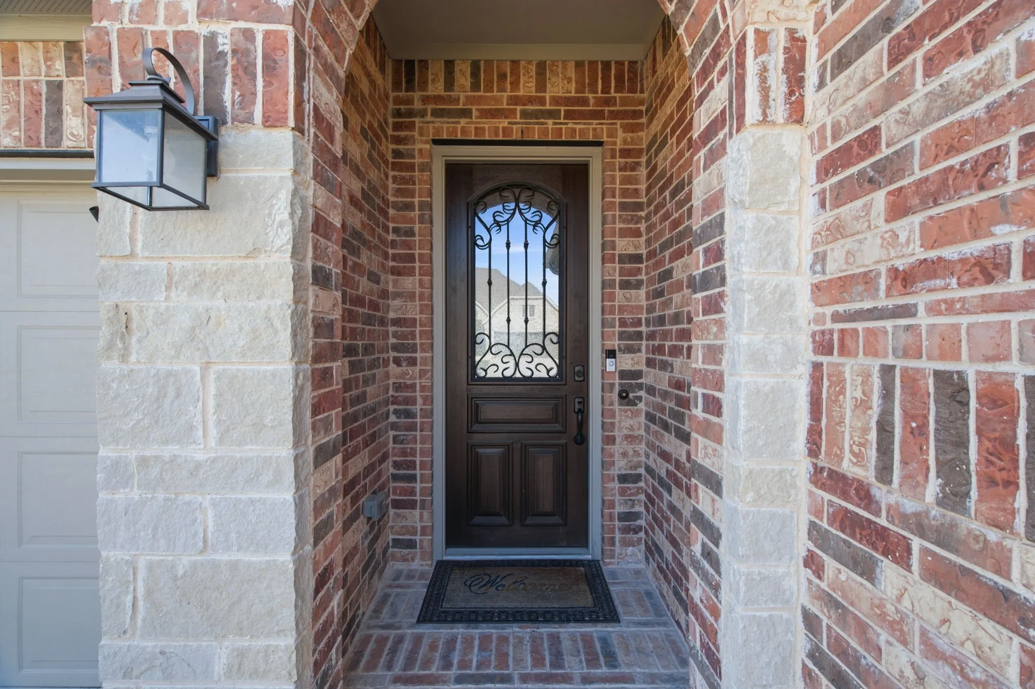 Arched entryway featuring a dark wood-finish door with decorative wrought iron detailing, brick and stone exterior, and an exterior wall lantern