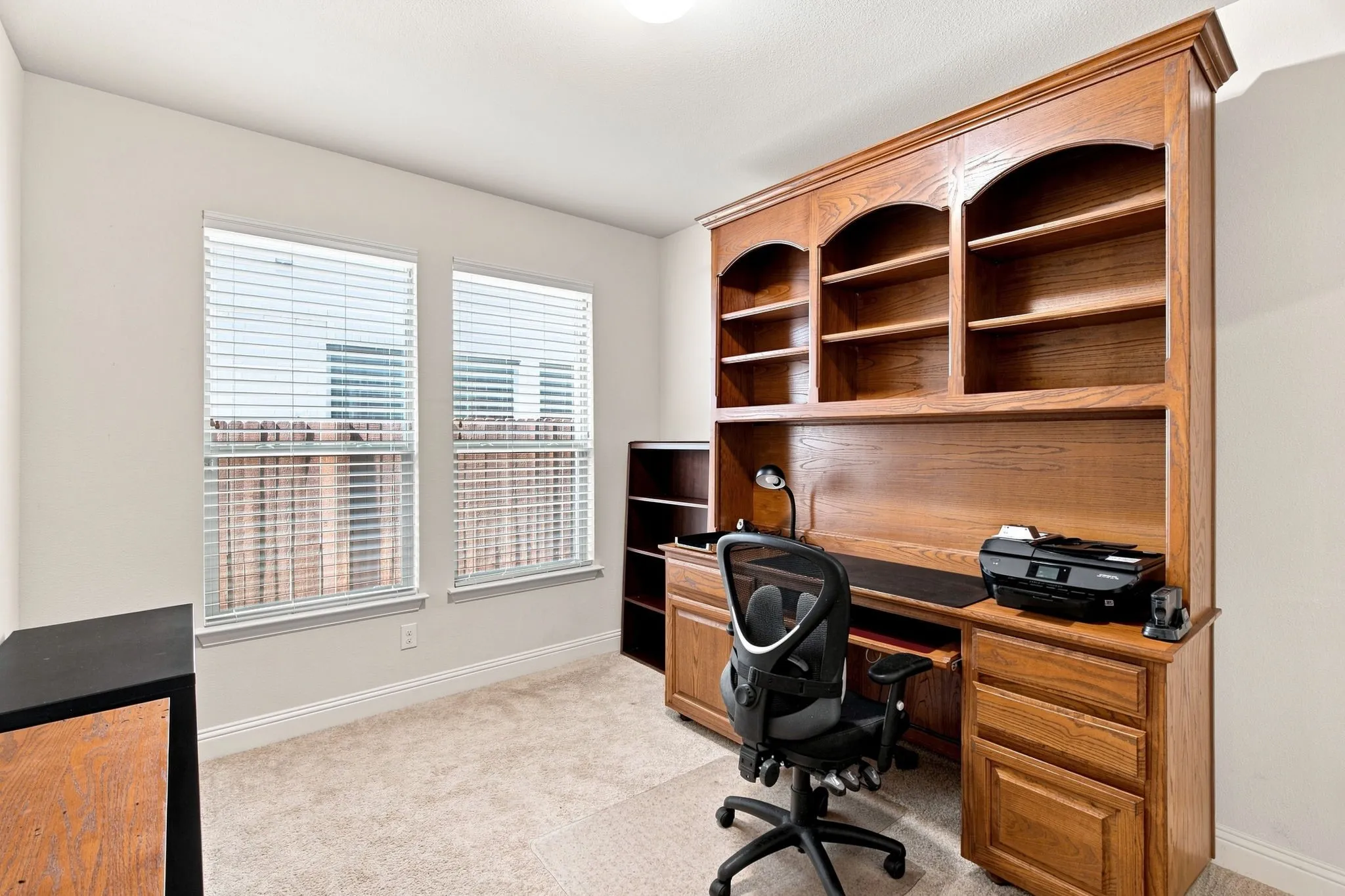 Built-in wood-finish shelving and desk system, neutral carpet flooring, and dual windows with blinds