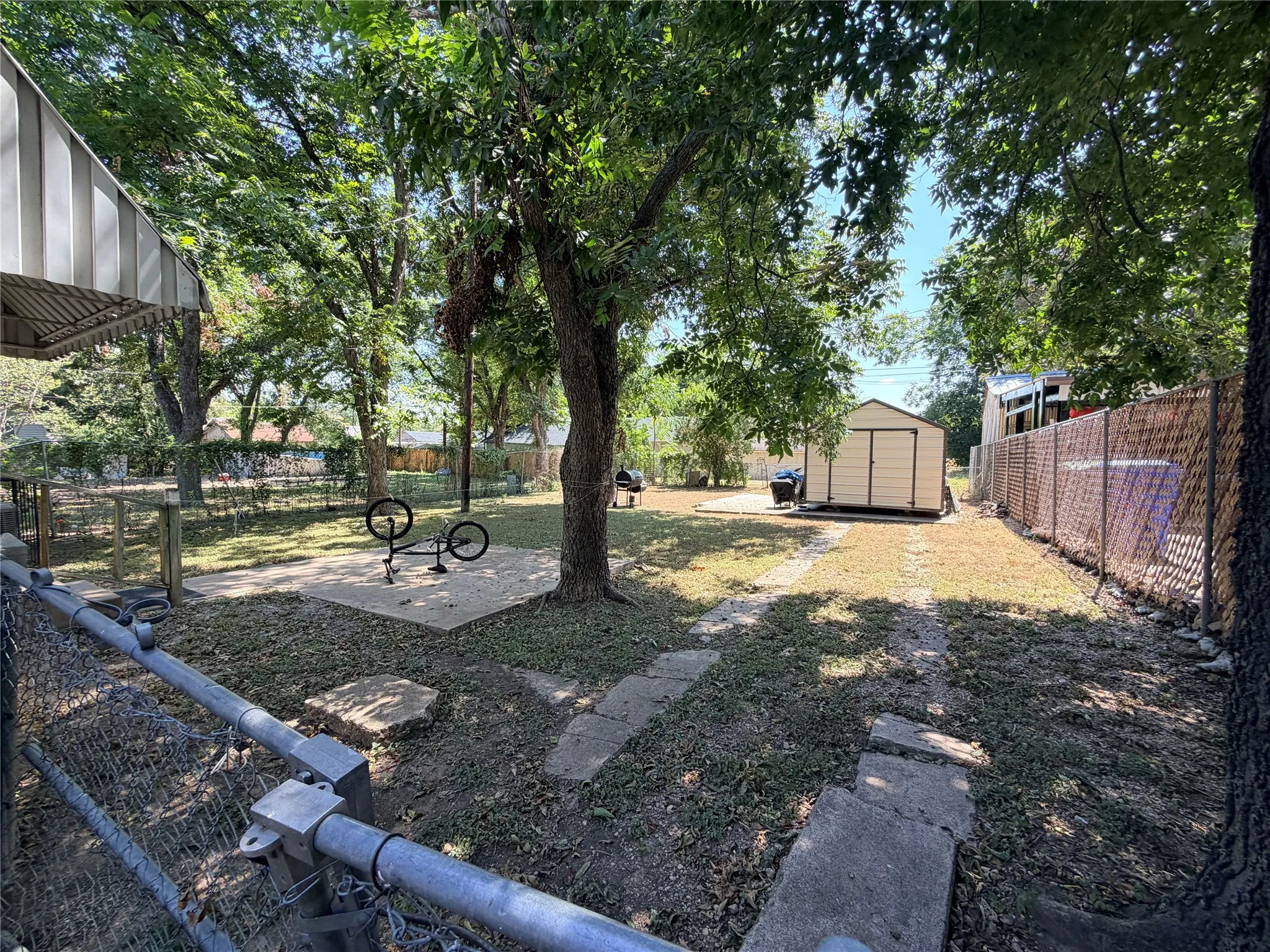 Expansive yard with mature trees, a detached storage shed, a paved patio area, and chain-link fencing