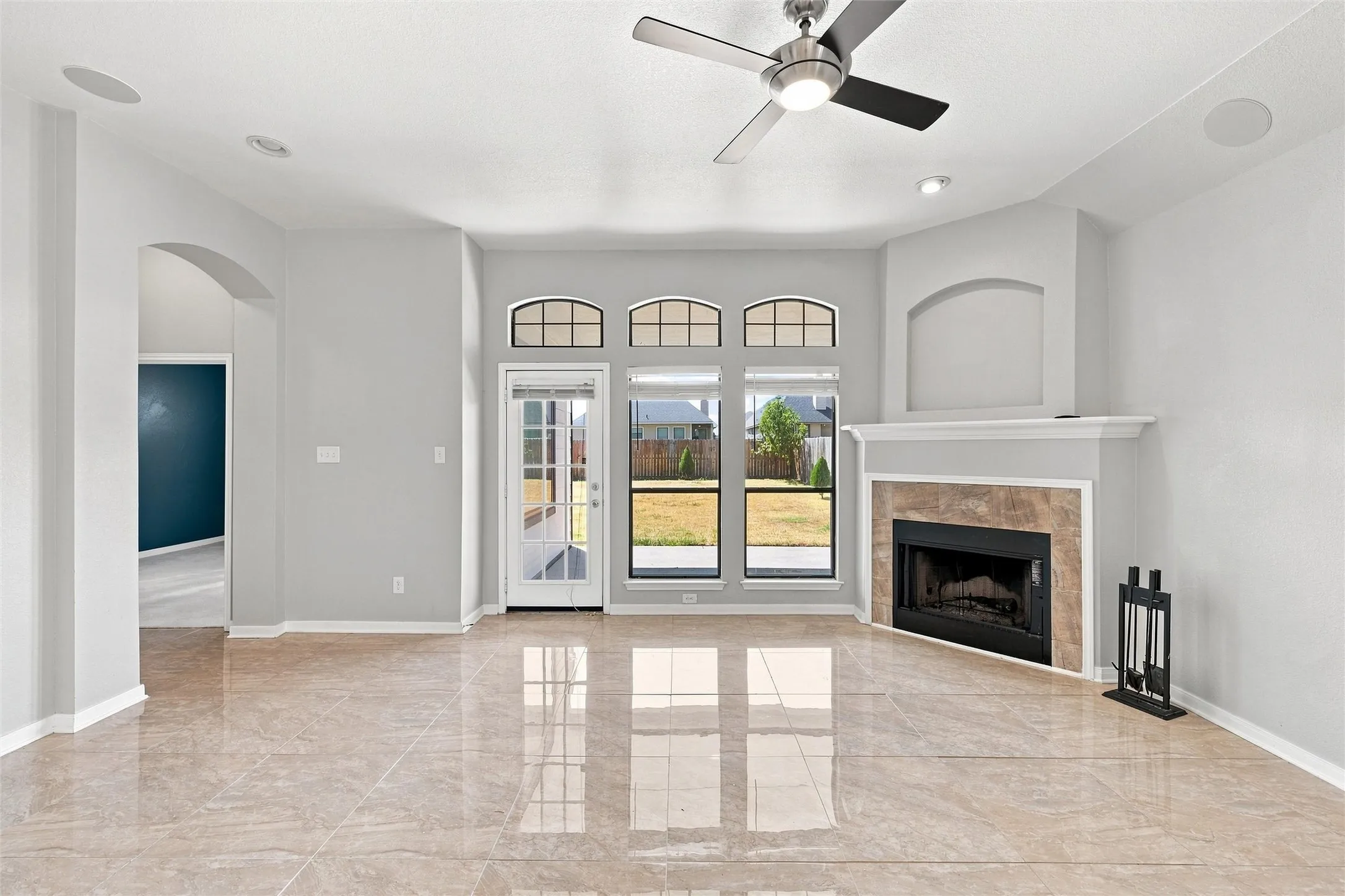 Bright open living area featuring polished stone-finish flooring, expansive French doors with transom windows opening to outdoor space, a wood-burning fireplace with surround, ceiling fan, and generous natural light throughout