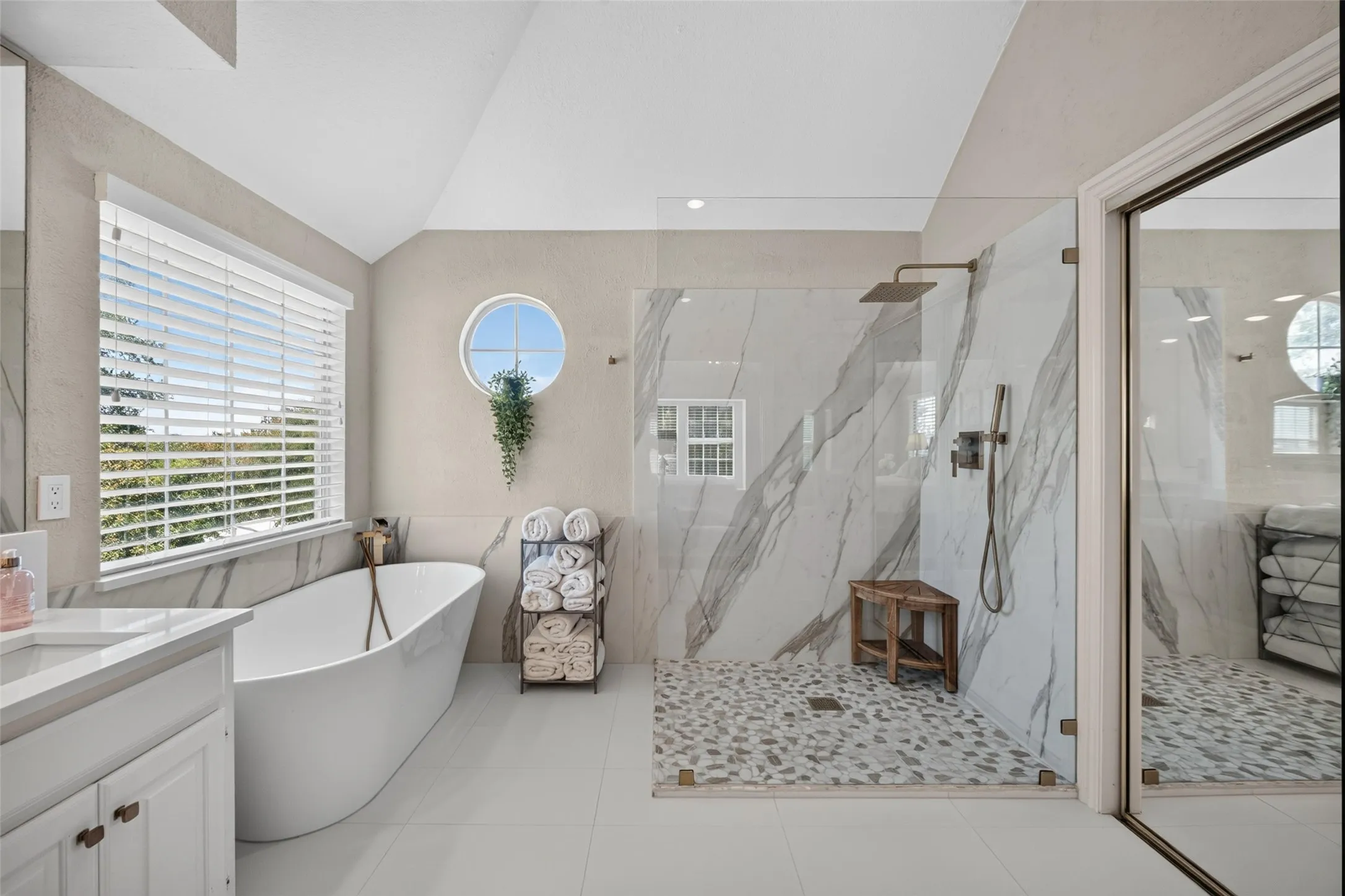Freestanding tub with floor-mounted faucet, large window with blinds, circular accent window, frameless glass shower enclosure, and pebble-tile shower floor