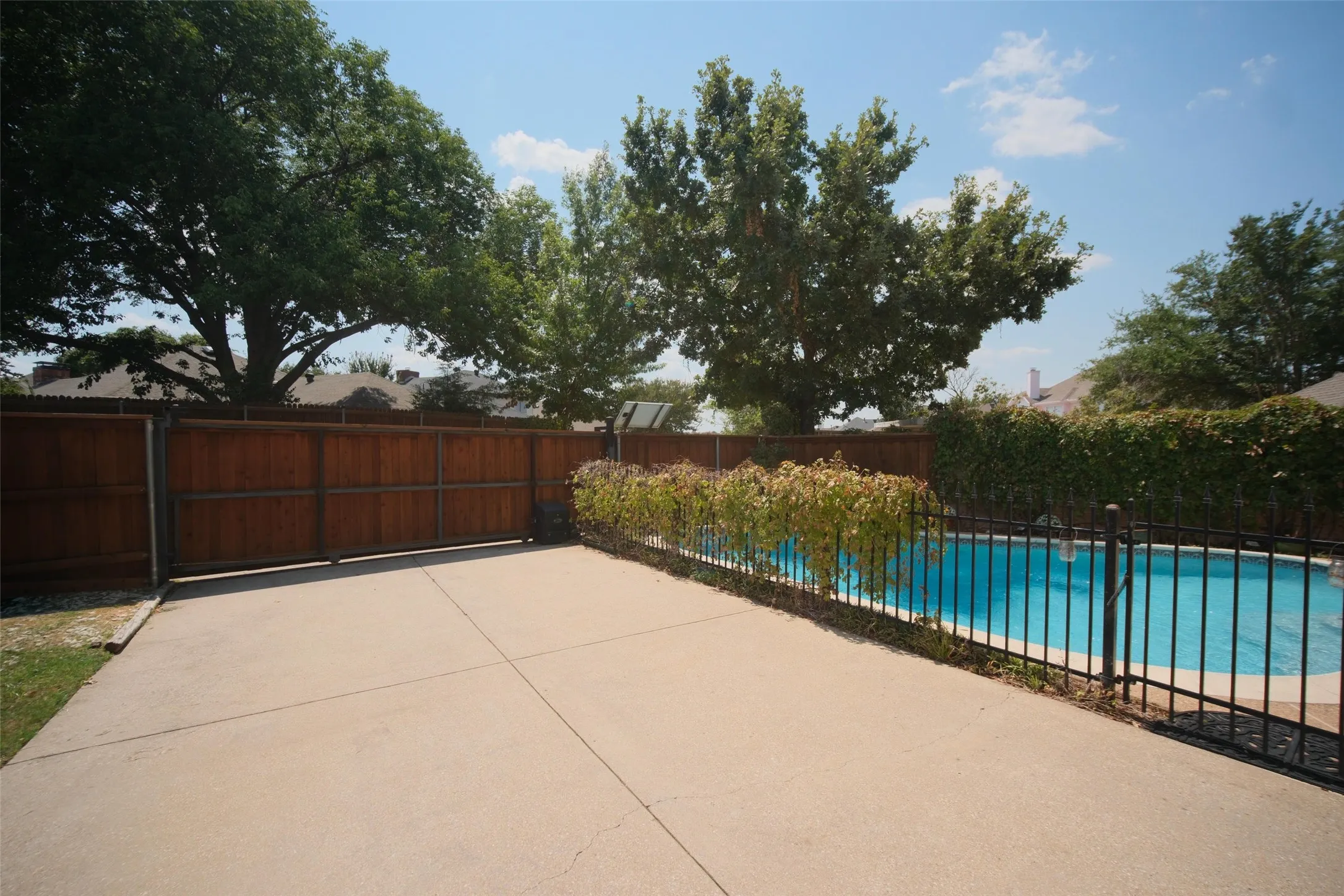 Swimming pool with security fencing, concrete patio area, mature trees, and a wood privacy fence