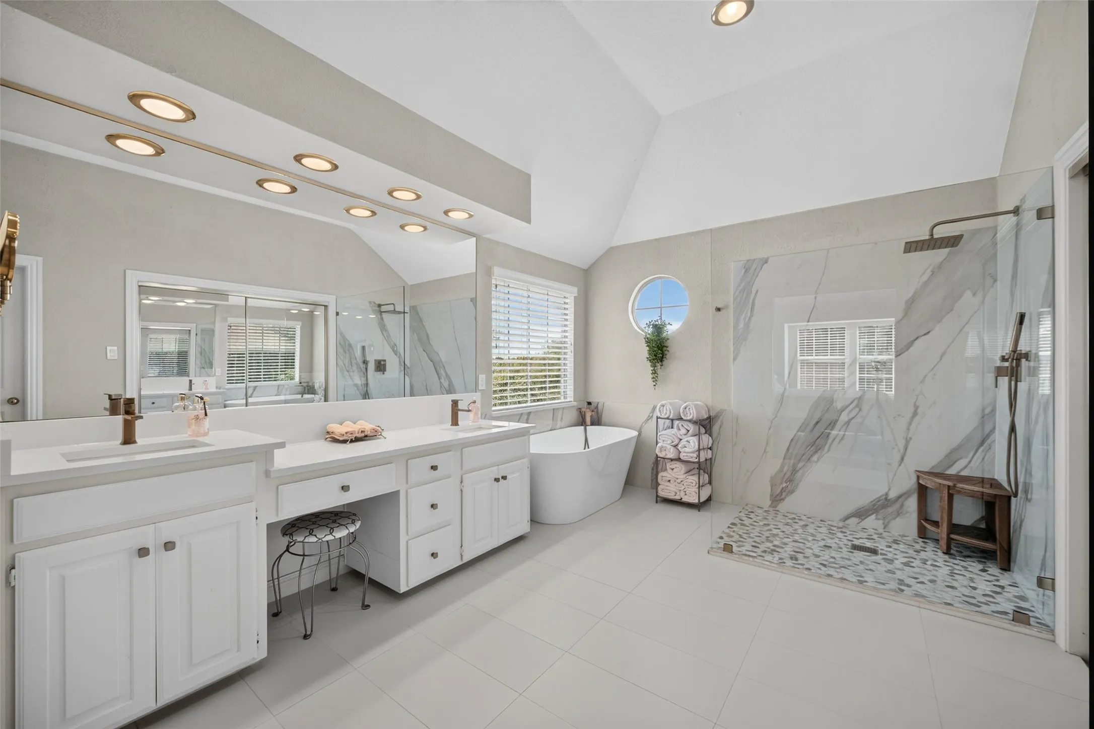 Vaulted ceiling bathroom featuring a dual vanity, large mirror with recessed lighting, freestanding tub, and a glass-enclosed shower with patterned tile flooring