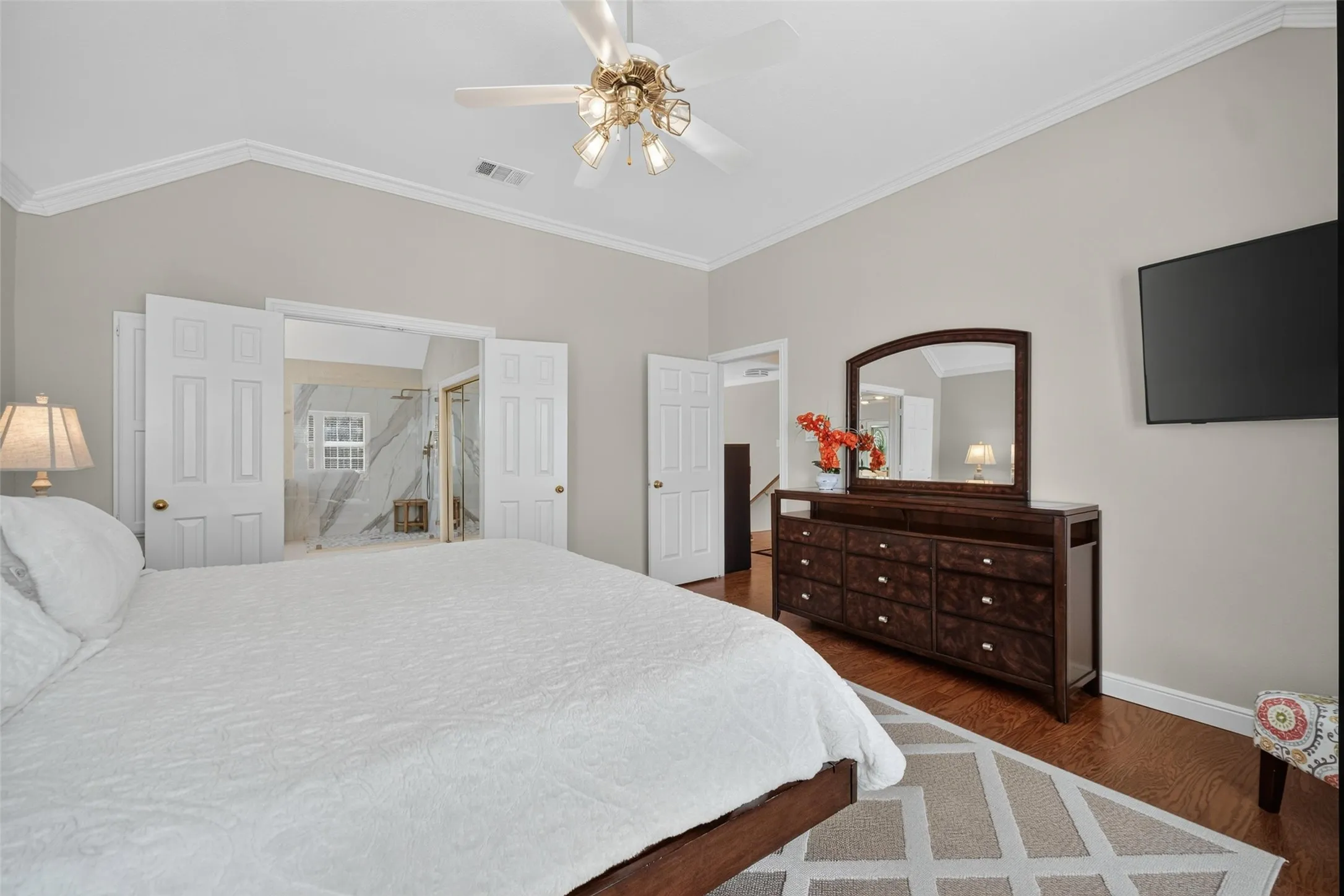 Vaulted ceiling with crown molding, ceiling fan, wood-finish flooring, and double doors opening to an ensuite bathroom with a glass-enclosed shower featuring tile finishes