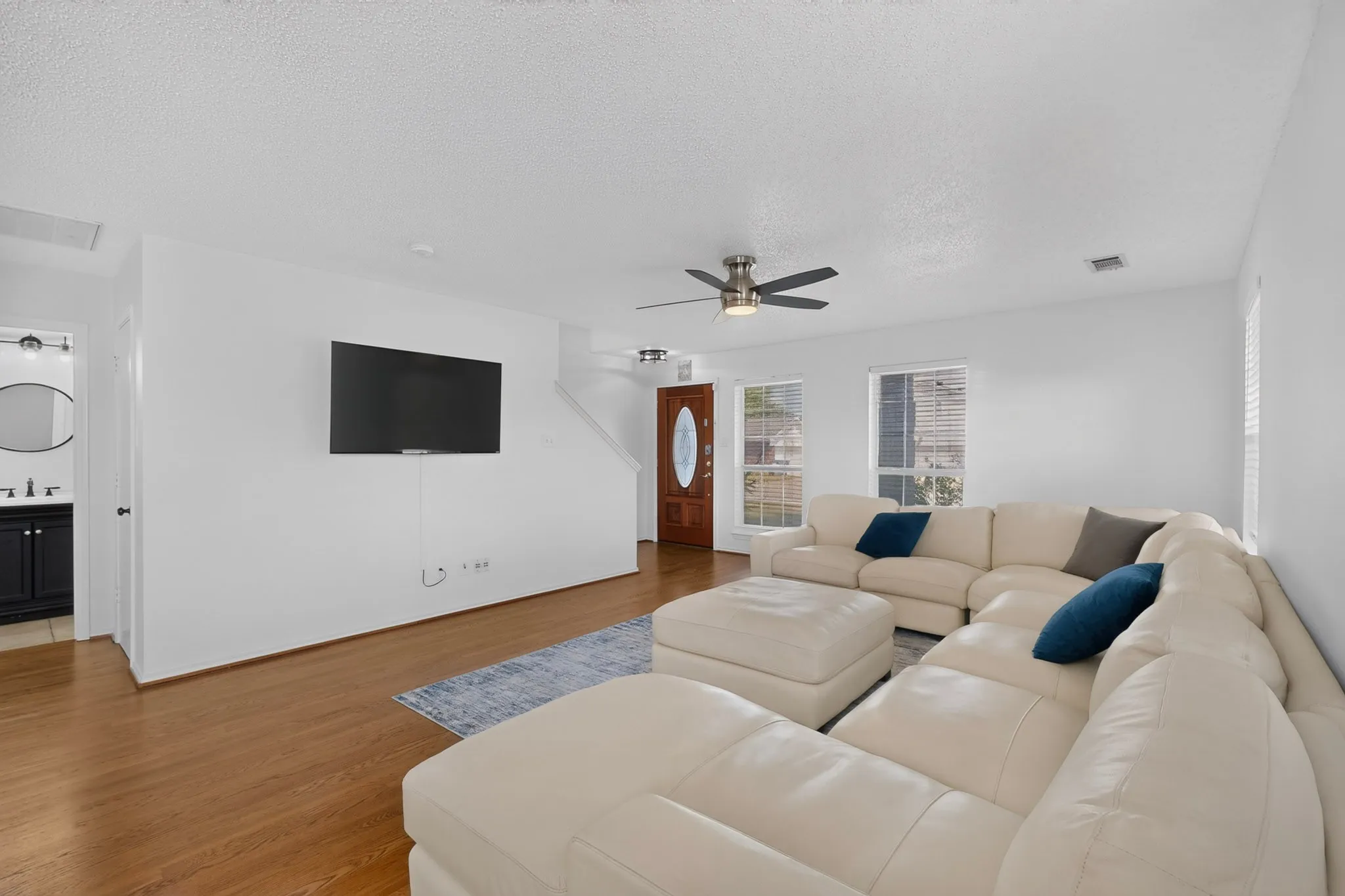 Wood-finish flooring, white walls, multiple windows with blinds, a ceiling fan with integrated lighting, and a wood-panel entry door with an oval glass insert