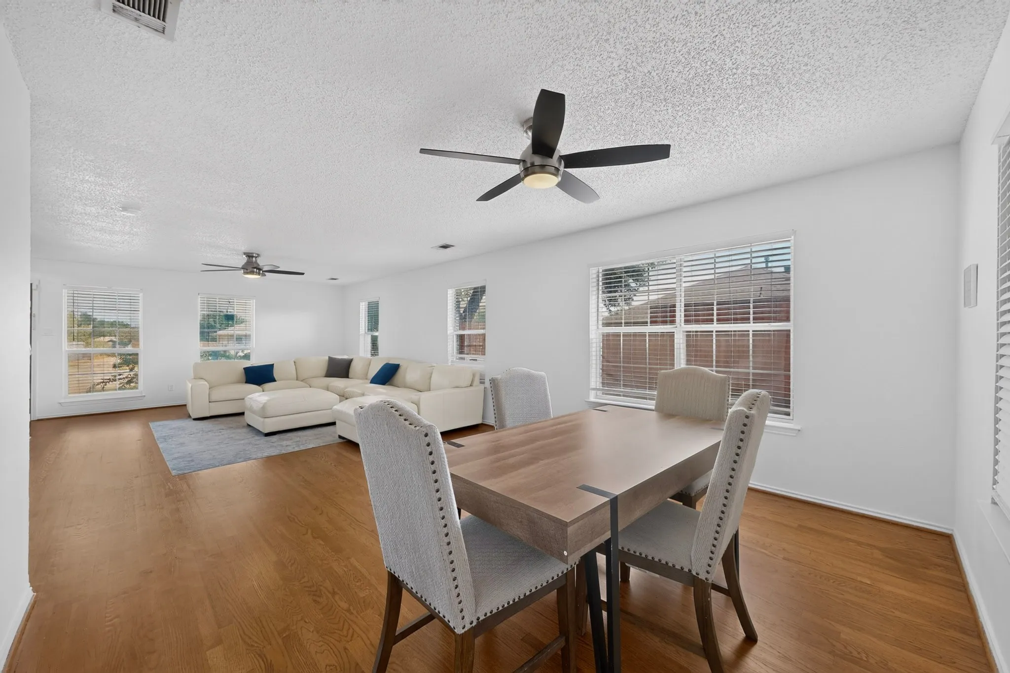 Expansive open-concept living area featuring wood-finish flooring, multiple windows with blinds, and two ceiling fans with integrated lighting