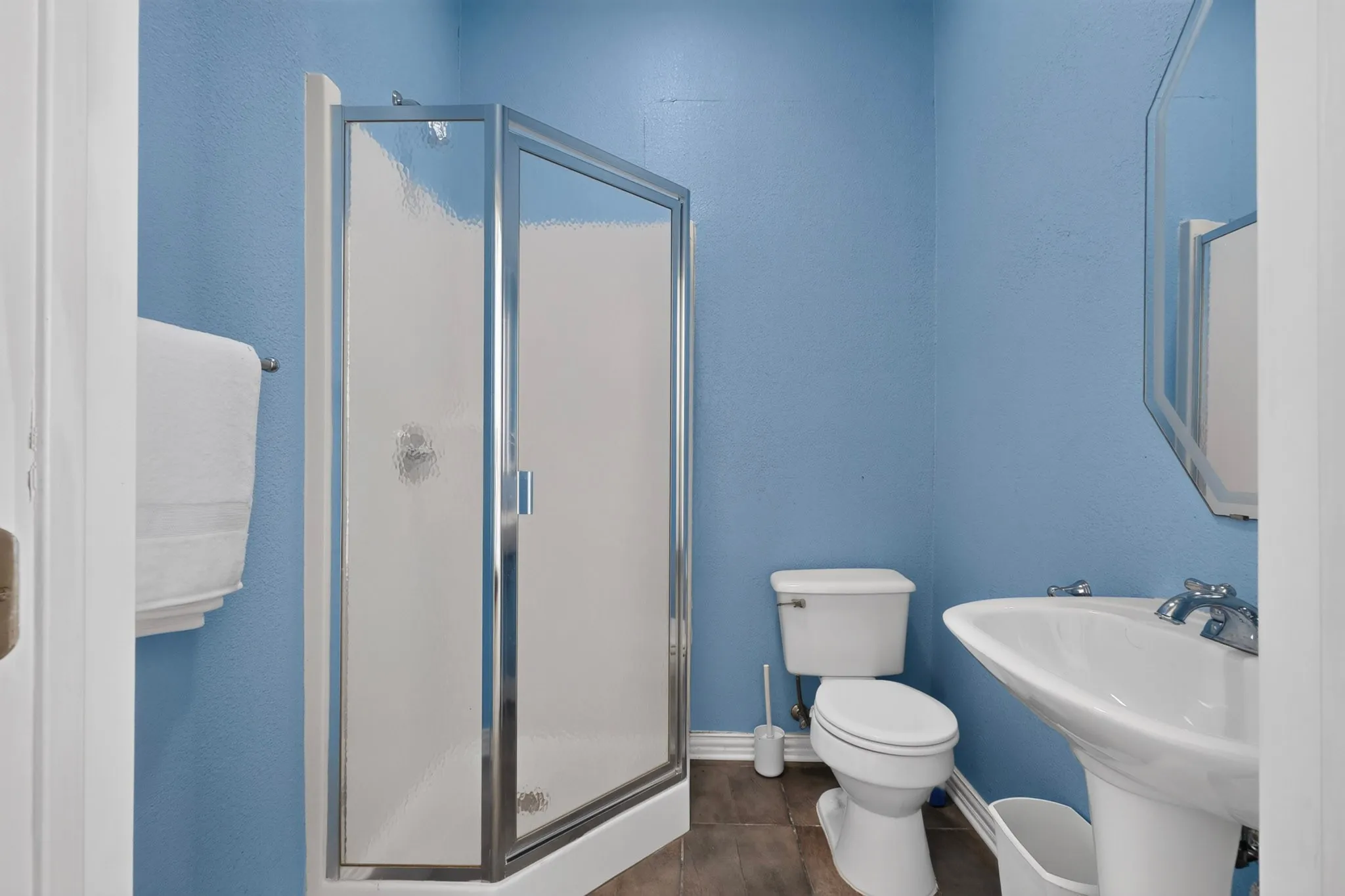 Corner shower with frosted glass enclosure, pedestal sink, wall-mounted mirror, and wood-finish flooring