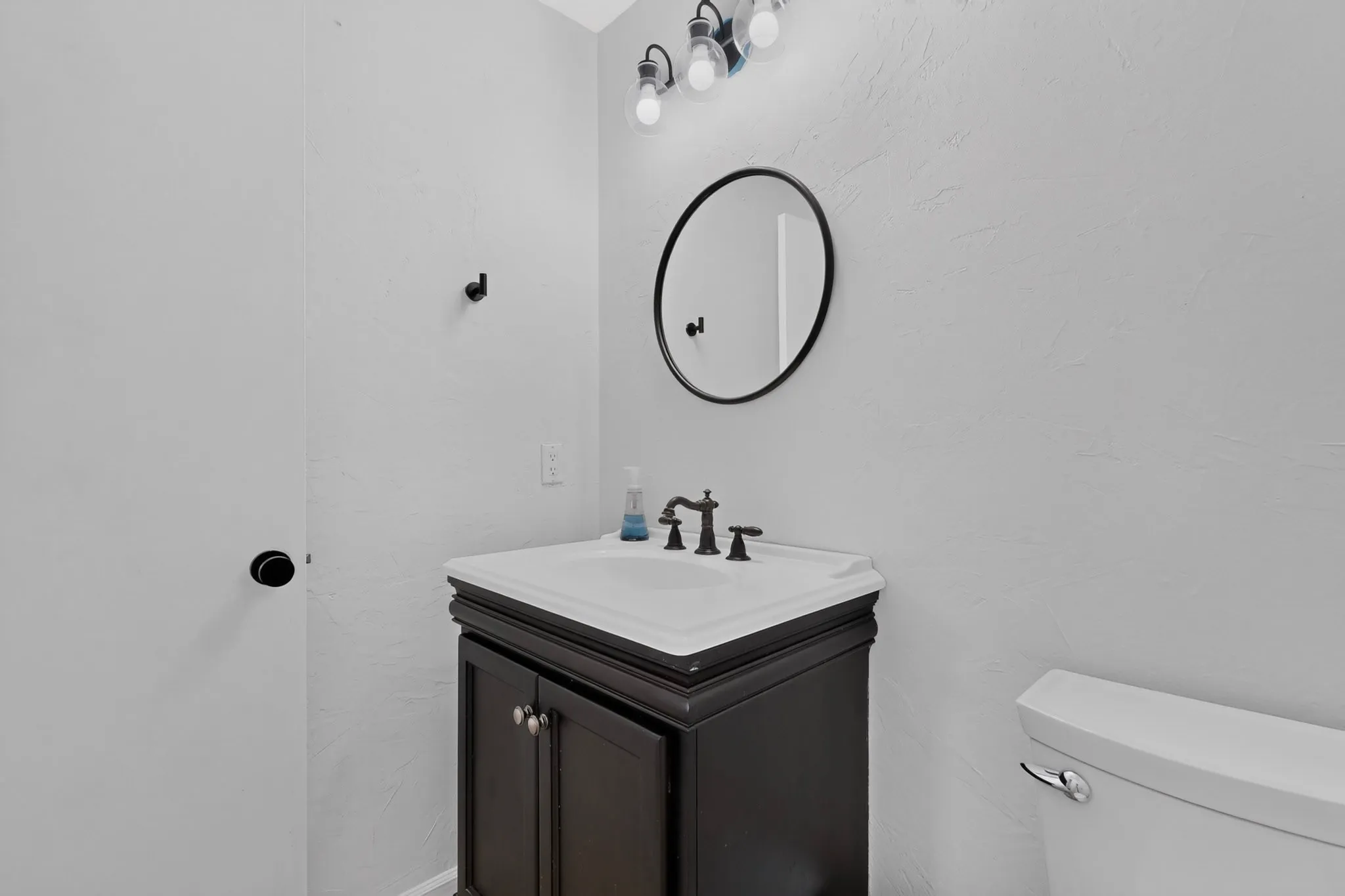 Vanity with a white basin and dark wood-finish cabinetry, circular mirror with a black frame, and black-finish wall-mounted lighting