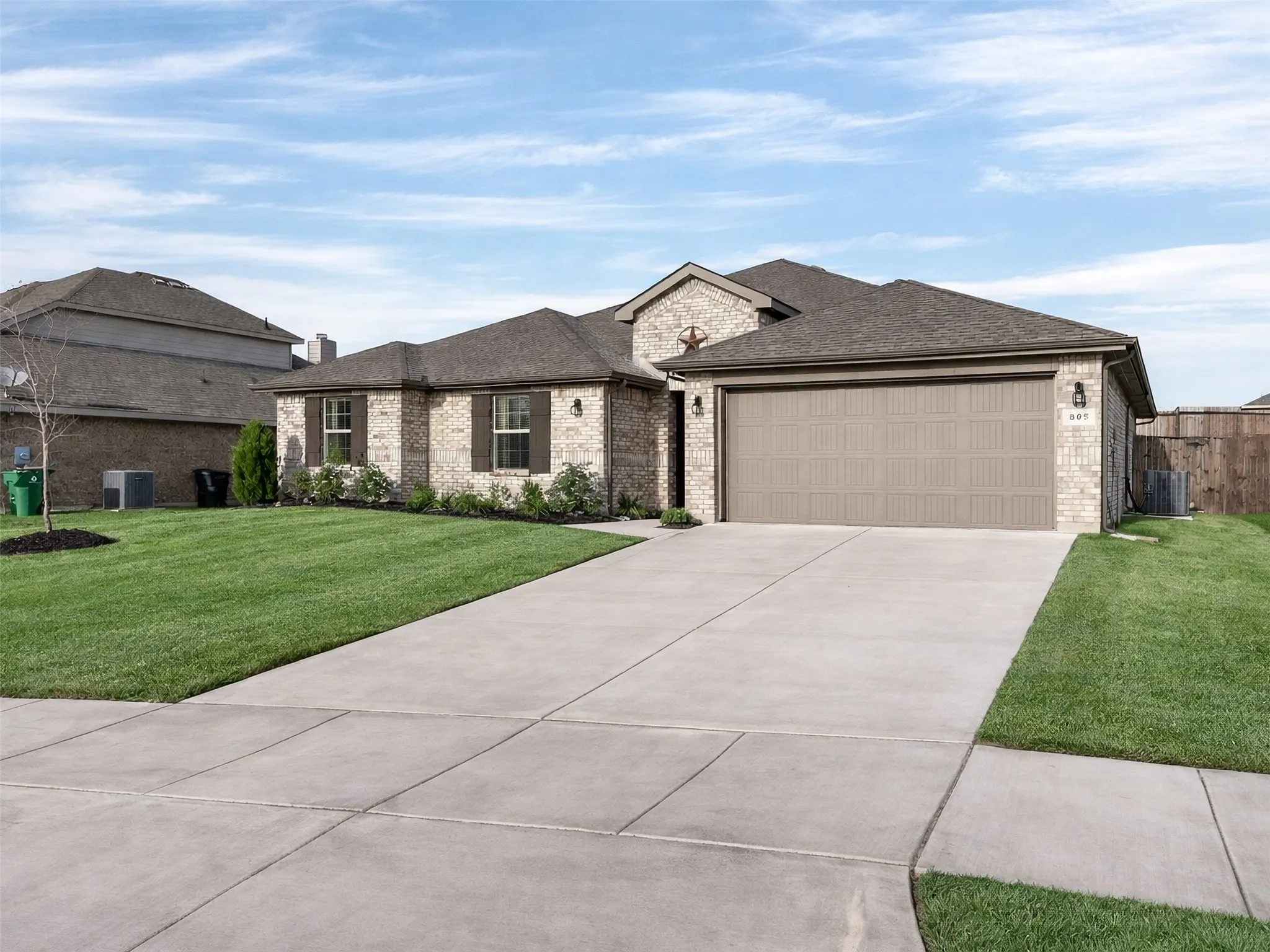 Brick exterior with dark shutters, attached garage, concrete driveway, and established front landscaping