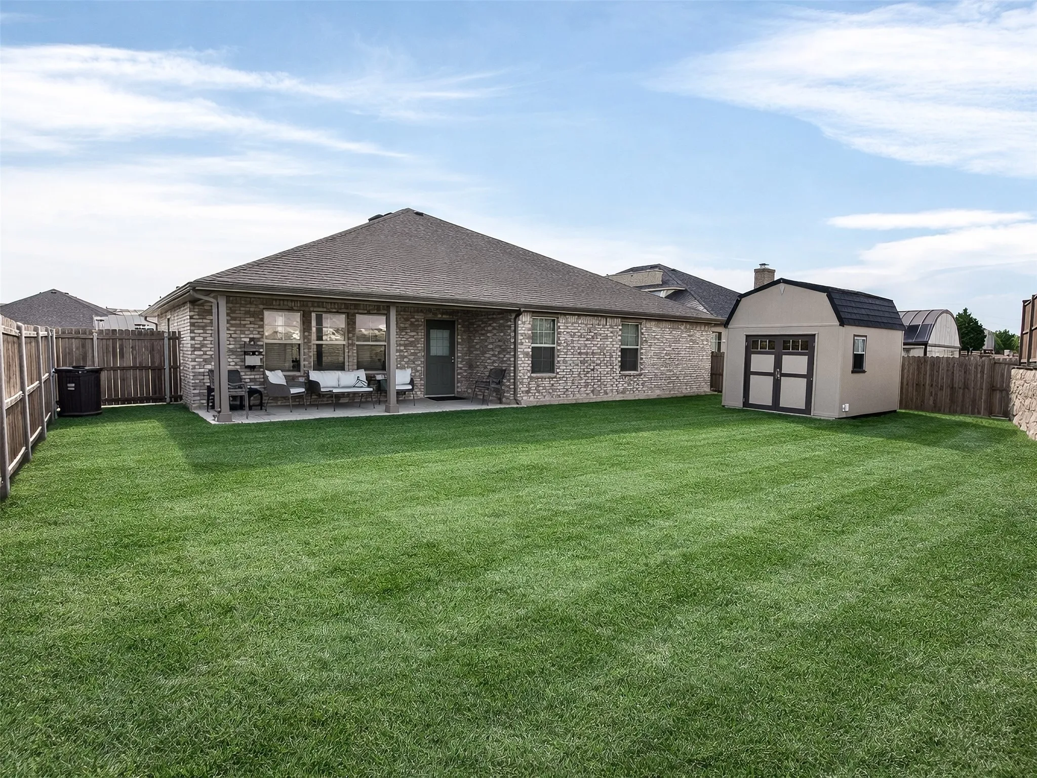 Expansive green lawn with a privacy fence, covered patio, brick exterior, and a detached storage shed