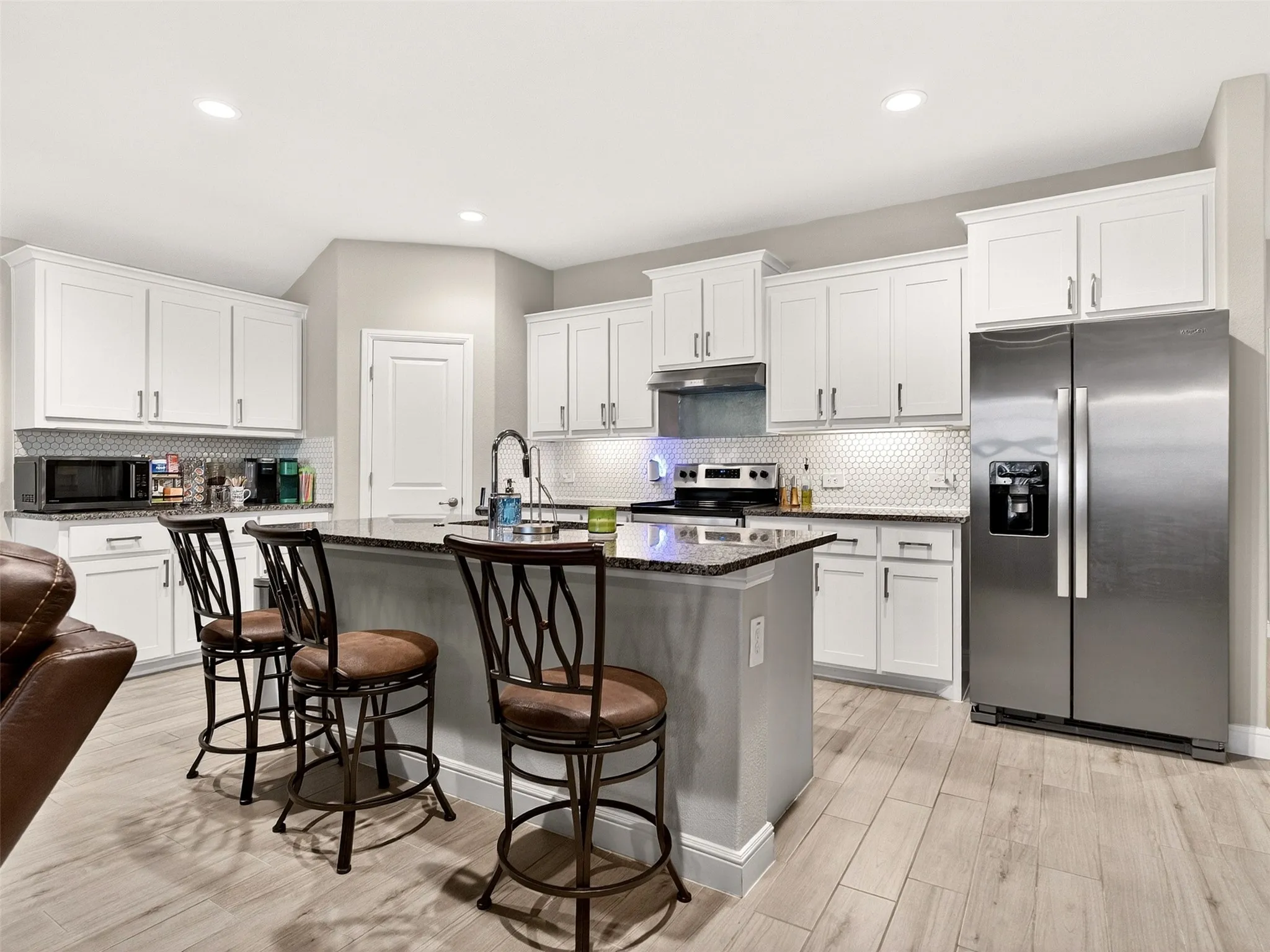 Kitchen featuring wood-finish flooring, an island with a dark stone-finish countertop, white shaker-style cabinetry, and a hexagonal tile backsplash