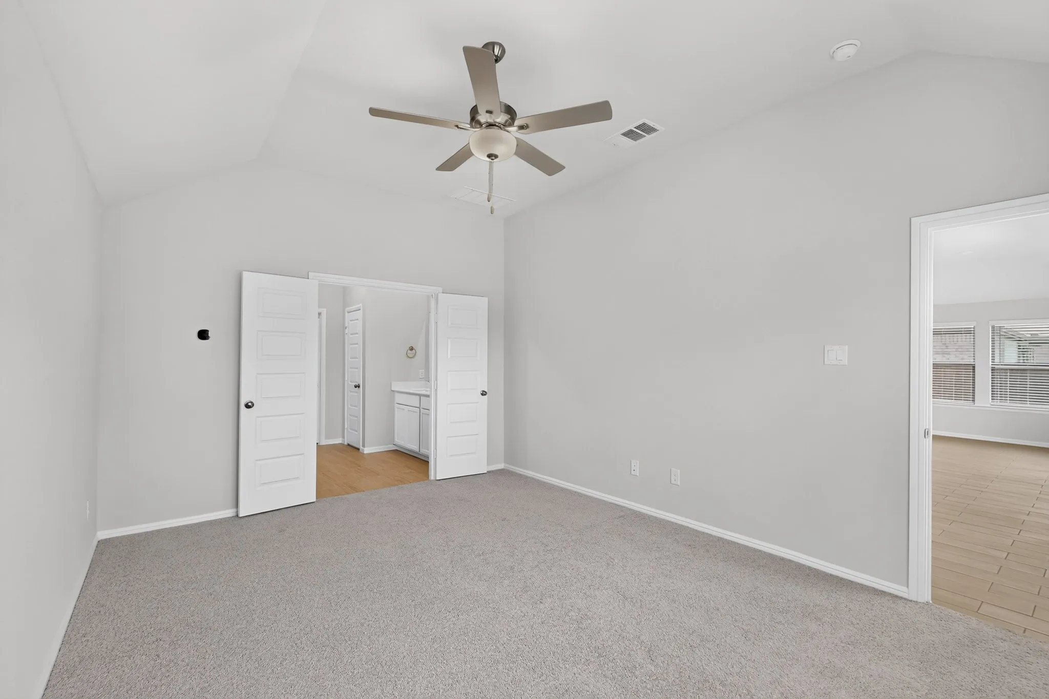 Vaulted ceiling with a ceiling fan, light gray carpeting, and white paneled doors leading to an adjoining space with wood-finish flooring and a vanity