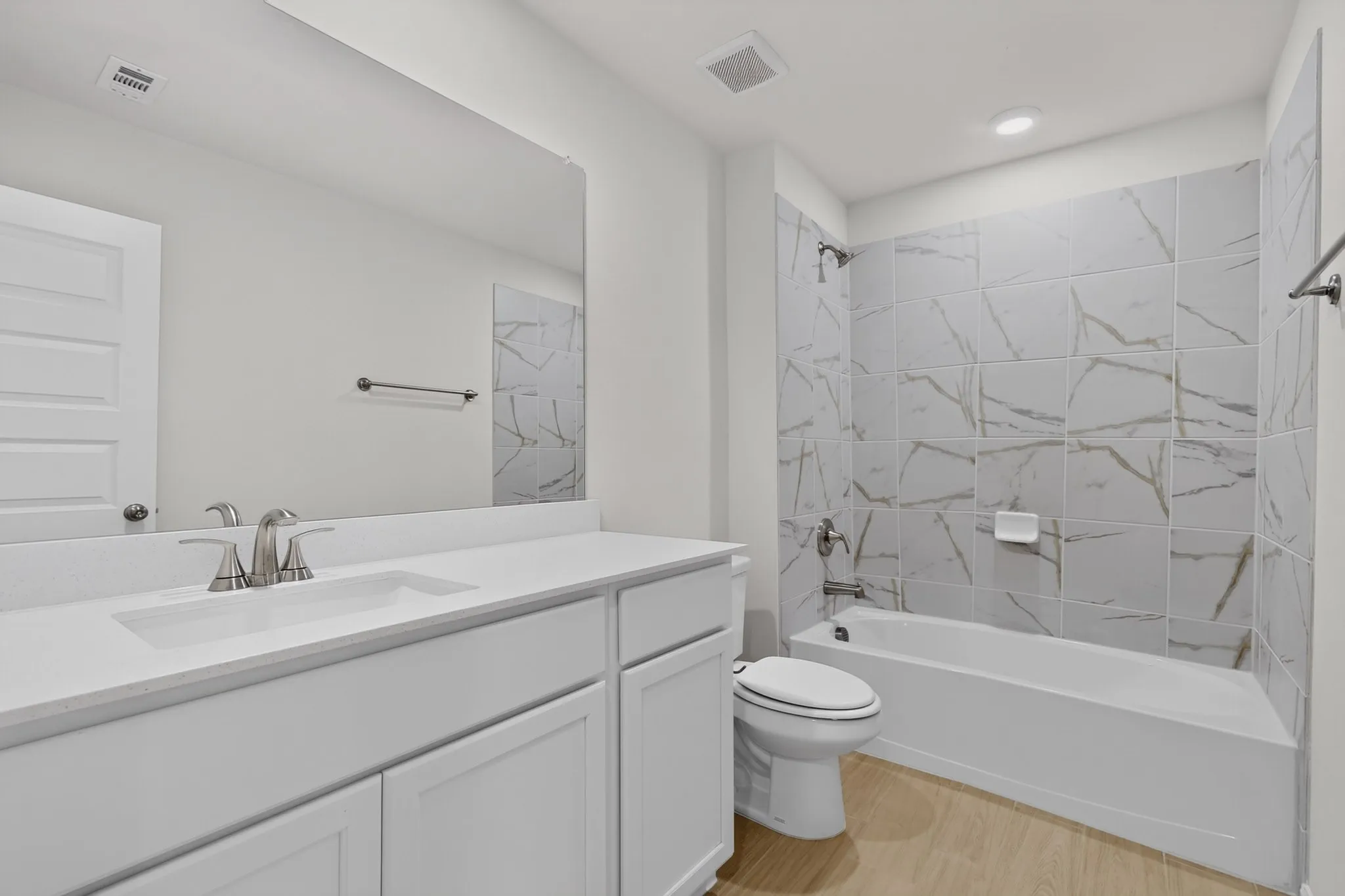Bathroom featuring a white vanity with an integrated sink, brushed nickel faucet, large wall mirror, bathtub with a tiled surround, and wood-finish flooring
