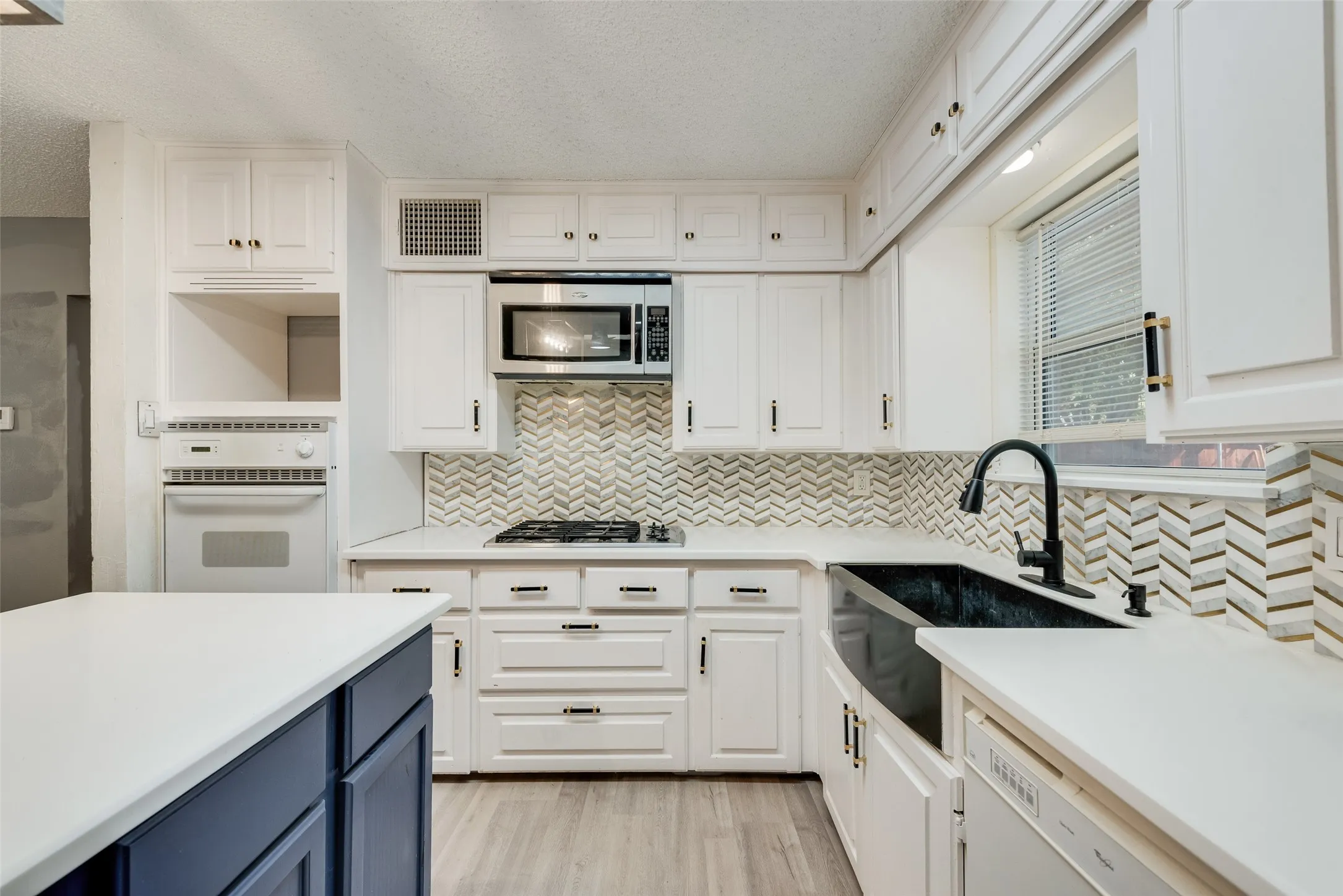 White cabinetry with dark hardware, herringbone tile backsplash, integrated stainless steel microwave, black farmhouse sink with a matte black faucet, and white countertops