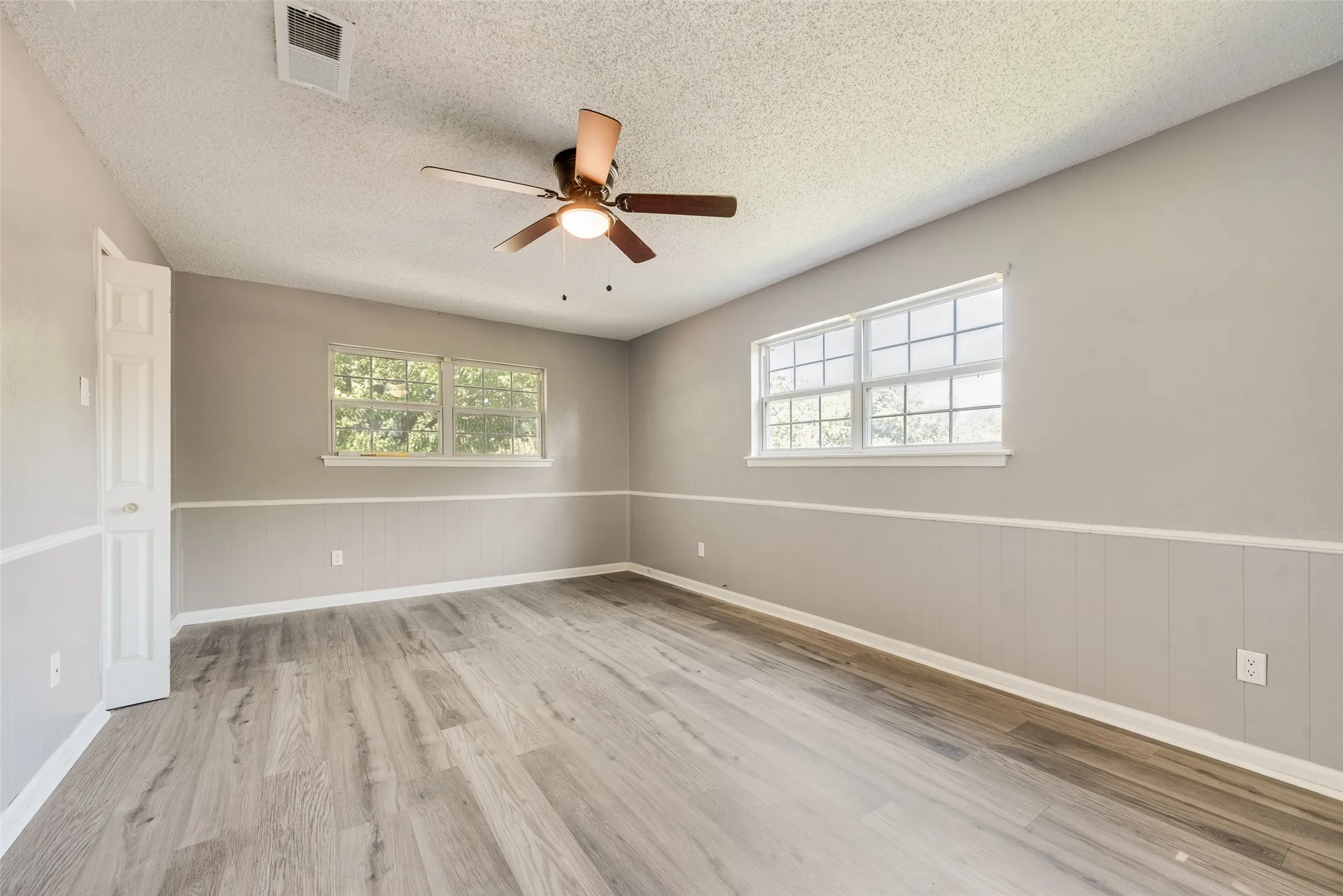 Spacious room featuring wood-finish flooring, two large windows, a ceiling fan, and wainscoting