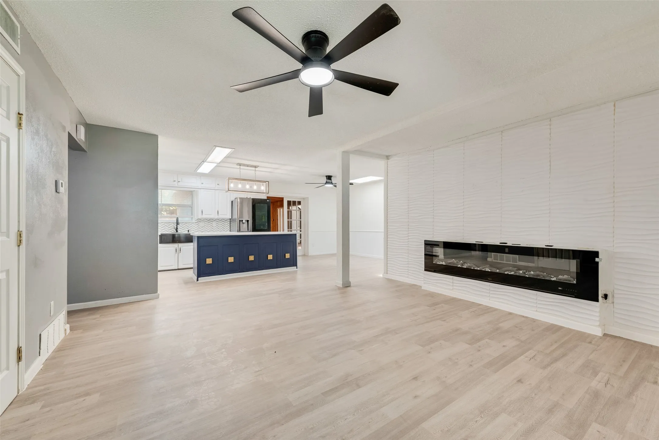 Open-concept living area with light wood-finish flooring, a contemporary electric fireplace, and a textured accent wall