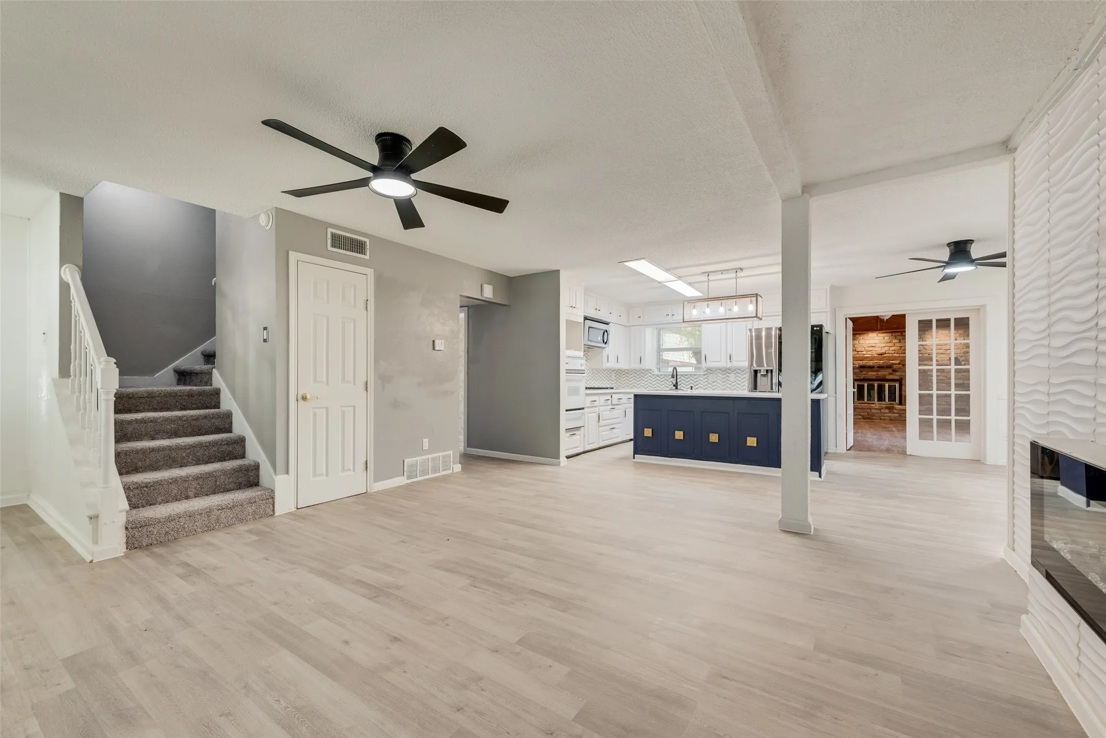 Wood-finish flooring, staircase with carpeted treads, modern kitchen island with contrasting cabinetry, built-in electric fireplace with textured surround, and French doors