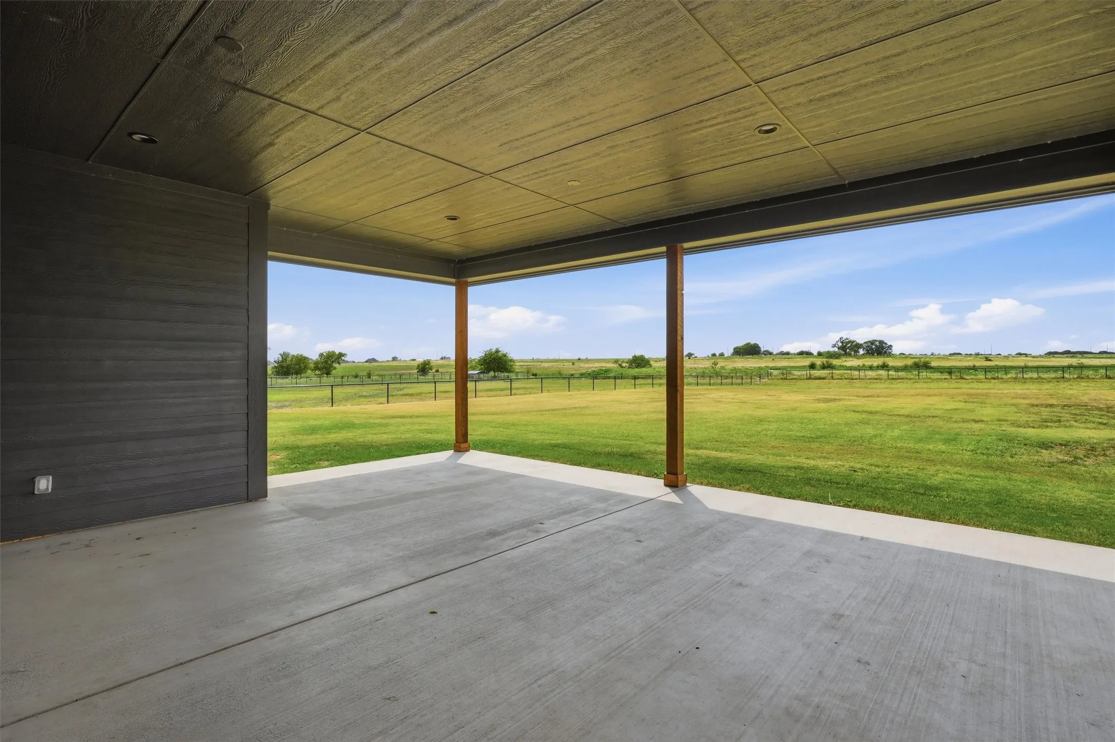 Expansive covered patio featuring a concrete surface, recessed lighting, and support columns, opening to a vast grassy expanse under a bright sky