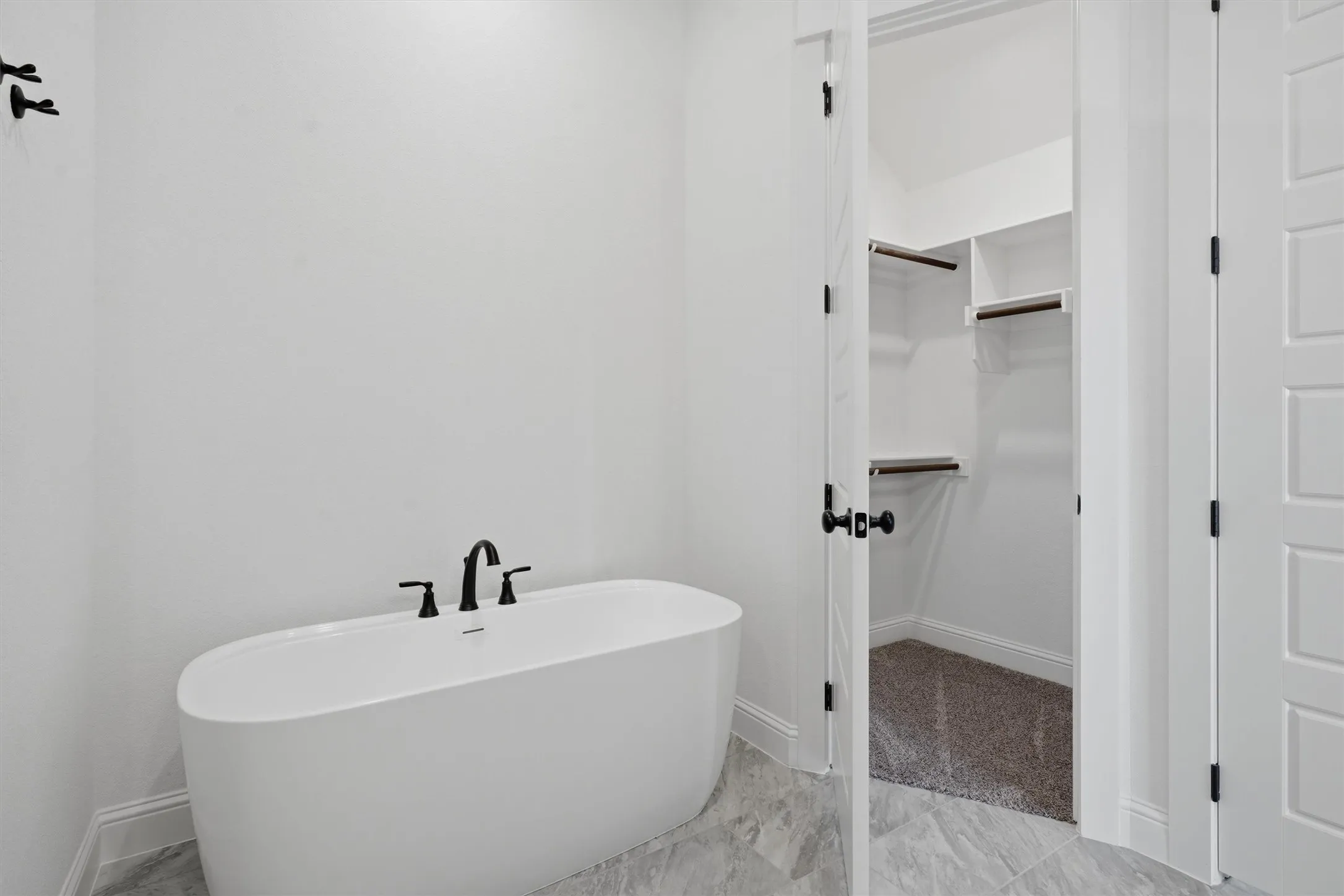 Freestanding soaking tub with matte black fixtures, gray patterned tile flooring, and an open closet with built-in shelving
