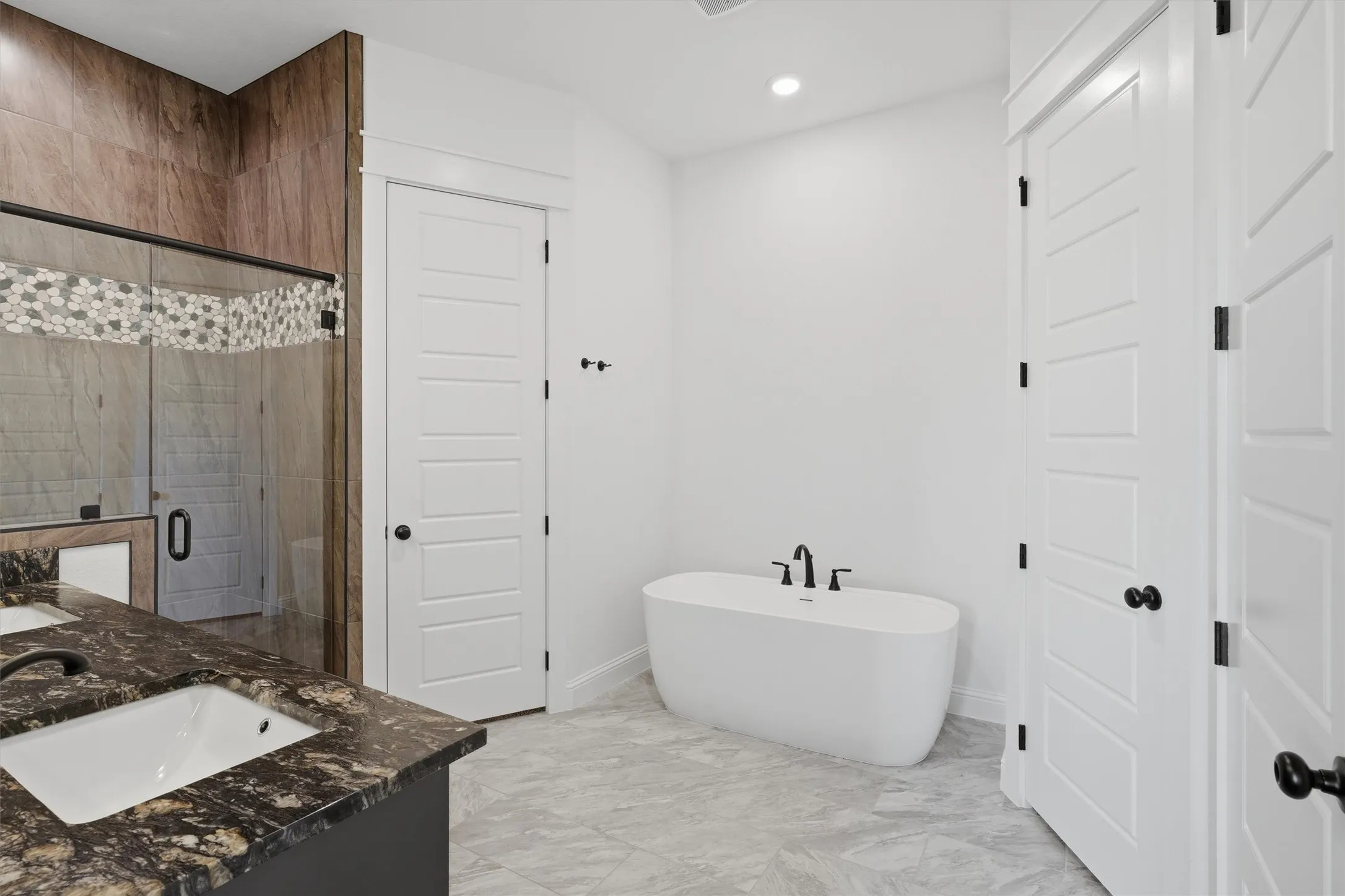 Freestanding soaking tub, dual vanity with undermount sinks and dark countertops, glass-enclosed shower with mosaic tile accent, and light-toned tile flooring