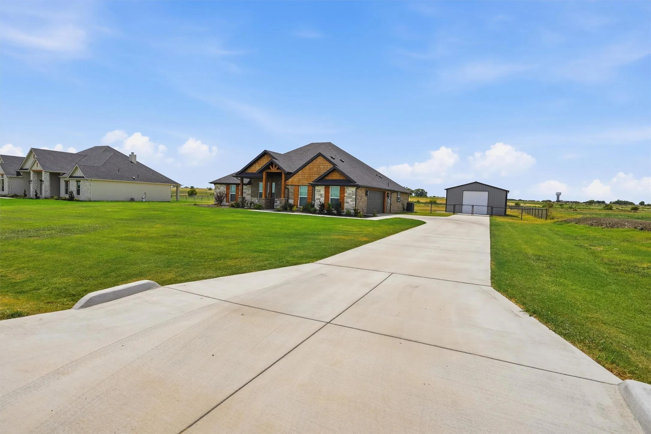 Expansive concrete driveway leading to a residence featuring stone and wood-look siding, a dark roof, and an auxiliary metal structure