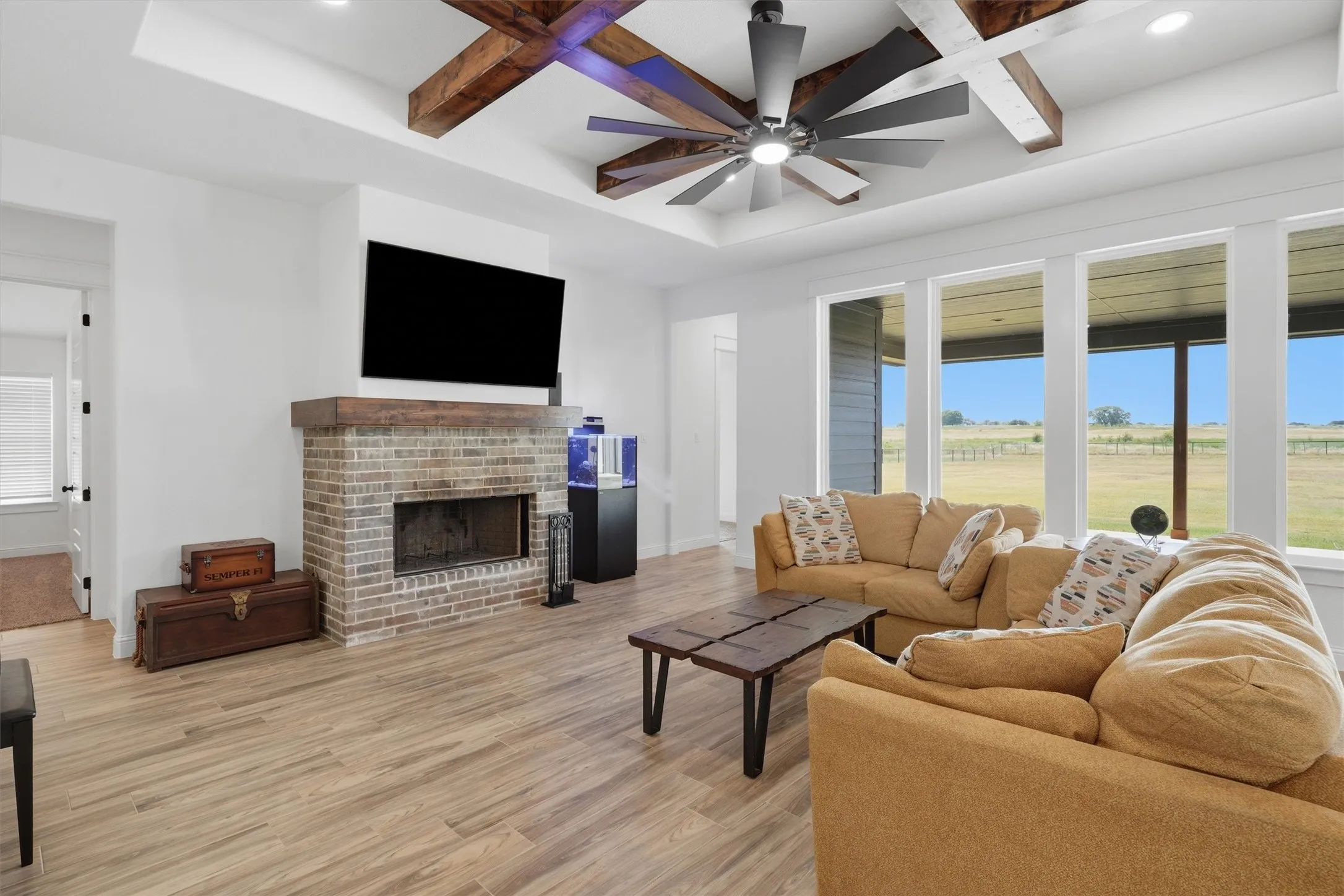 Living space featuring a brick fireplace with a wood mantle, wood-finish flooring, a tray ceiling with exposed wood beams, and an integrated ceiling fan