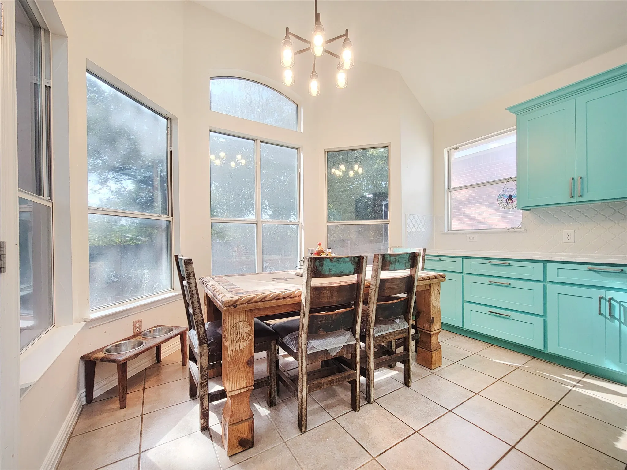 Vaulted ceiling with an architectural chandelier, expansive windows, turquoise cabinetry, white subway tile backsplash, and tile flooring