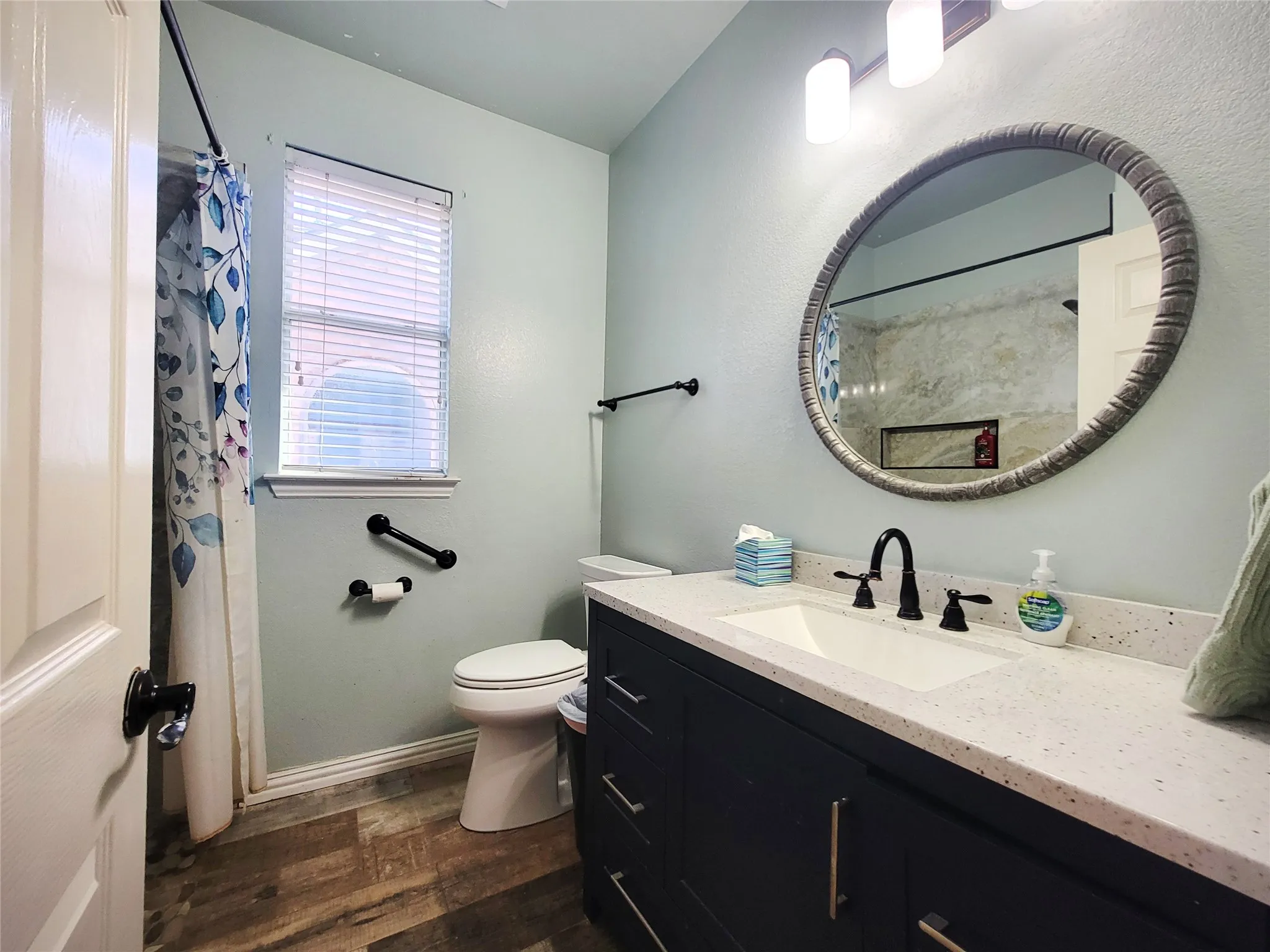Step in shower, Vanity with a white countertop and undermount sink, wood-finish flooring, oval mirror, and a window with blinds