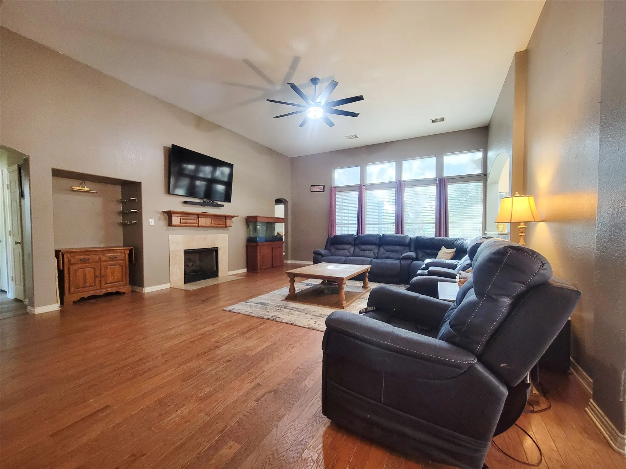 Vaulted ceilings with a ceiling fan, wood-finish flooring, recessed built-in shelving, fireplace with stone surround and wood mantle, and a wall of windows