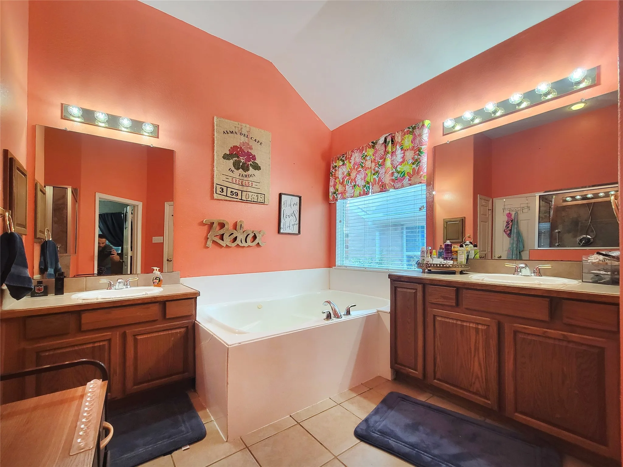Dual vanity sinks with wood cabinetry, a large soaking tub, tile flooring, and a vaulted ceiling, walk in closet