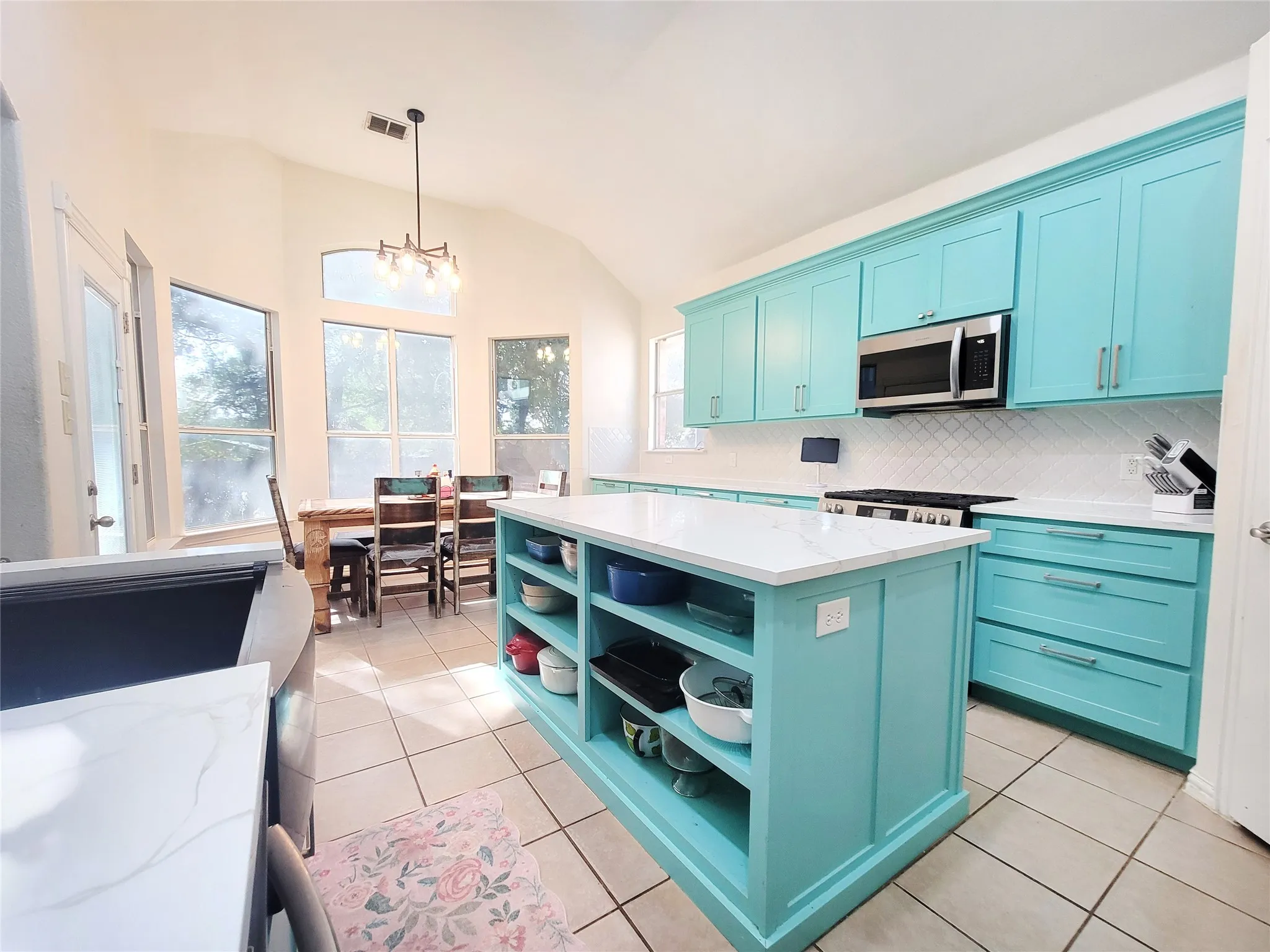 Kitchen featuring custom blue cabinetry, stainless steel appliances, a center island with open shelving, tile flooring, and vaulted ceilings with a contemporary light fixture