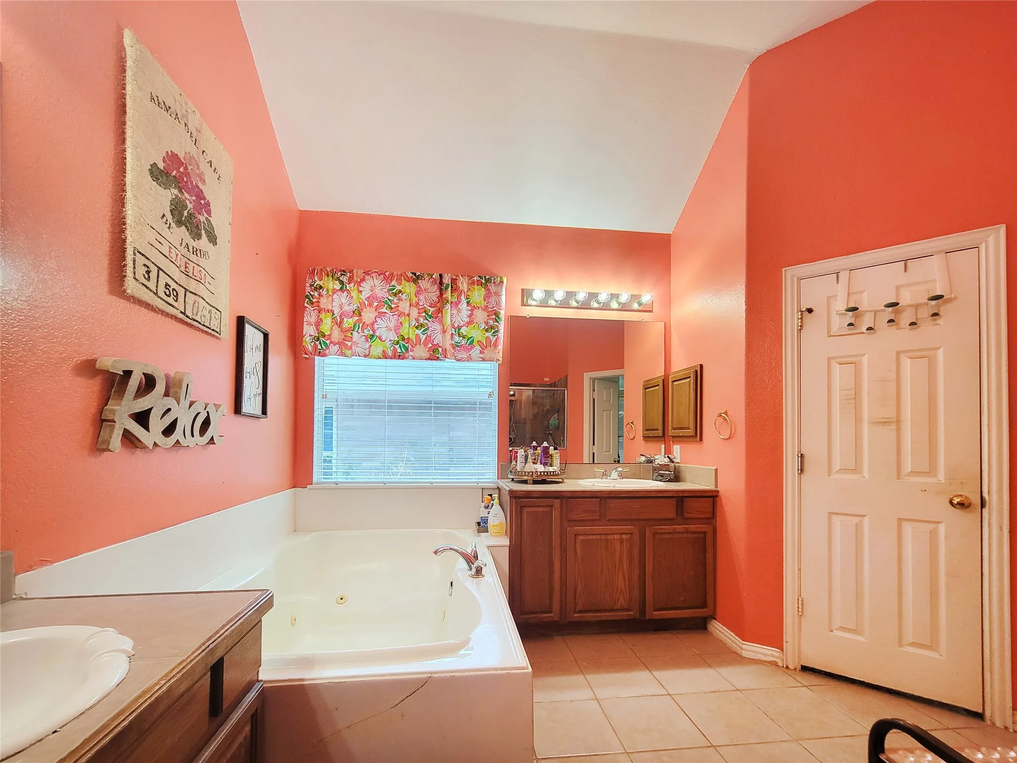 Vaulted ceiling, built-in jetted tub, wood-finish vanity with a single basin sink, large wall-mounted mirror, and tile flooring