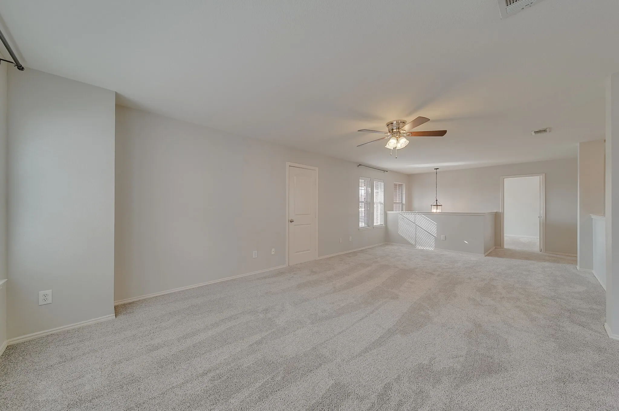 Expansive carpeted space featuring a ceiling fan with light fixture, multiple windows, interior doors, and a staircase with a railing