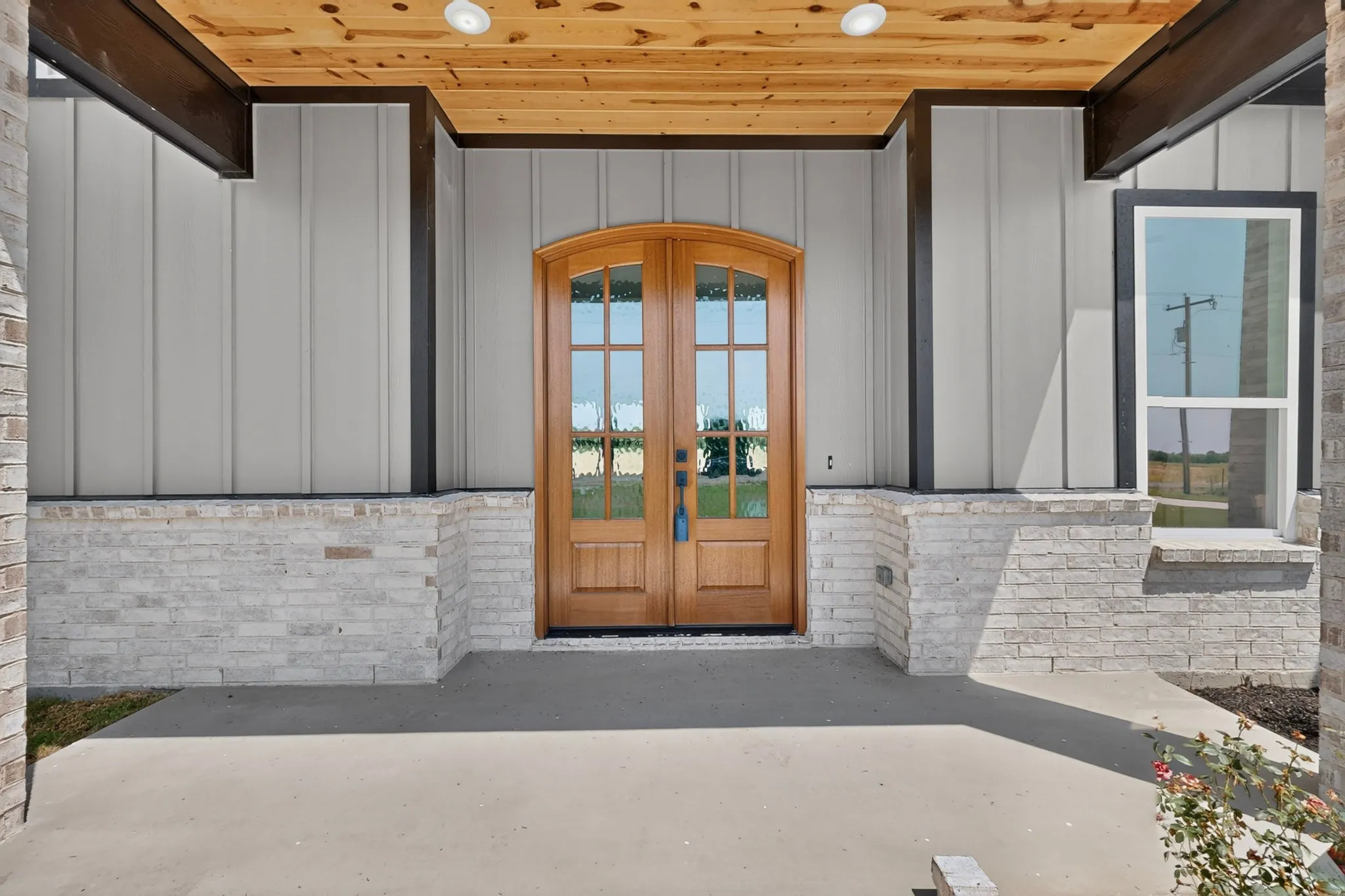 Arched wood-finish double entry doors, vertical panel siding, light-toned brick accents, concrete porch, and a wood-paneled ceiling with recessed lighting