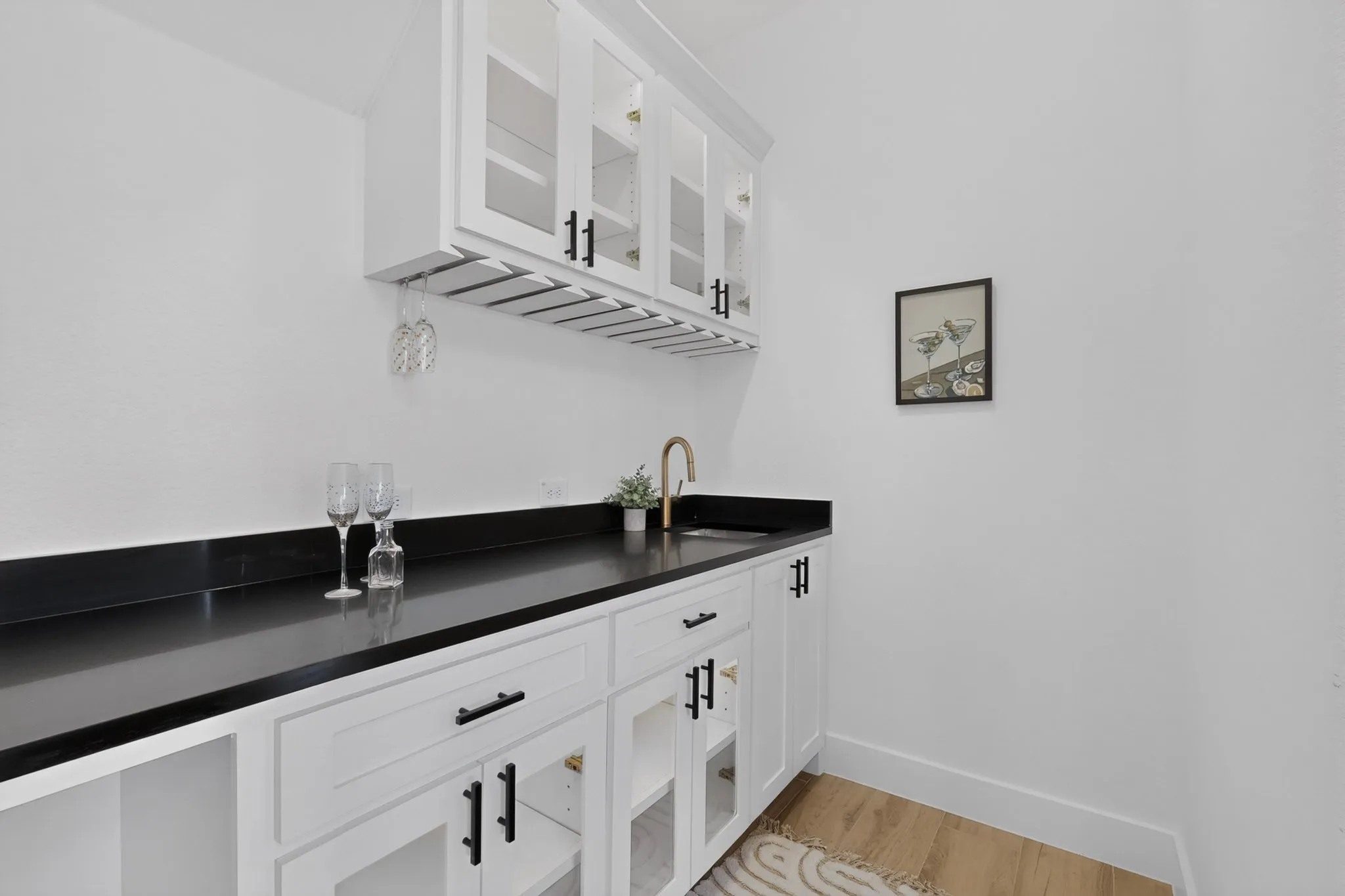 White cabinetry with black hardware, black countertops, undermount sink with brass-finish faucet, upper glass-front cabinets, and wood-finish flooring