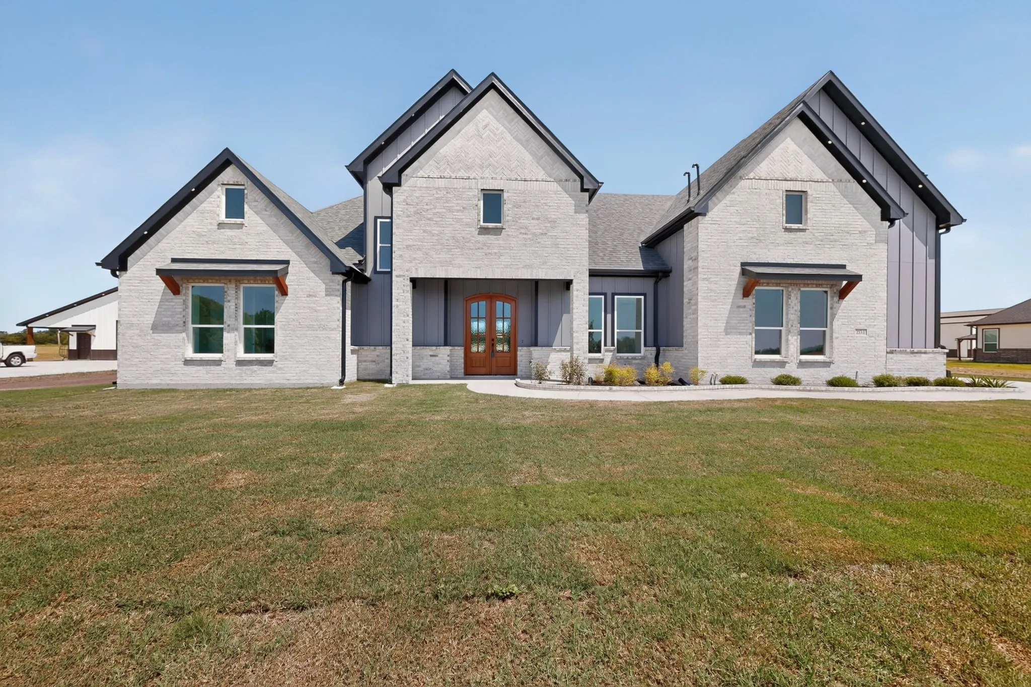 Modern exterior design featuring light-colored brick, dark-toned vertical siding, a wood-finish double-door entry, a gabled roofline, and established landscaping