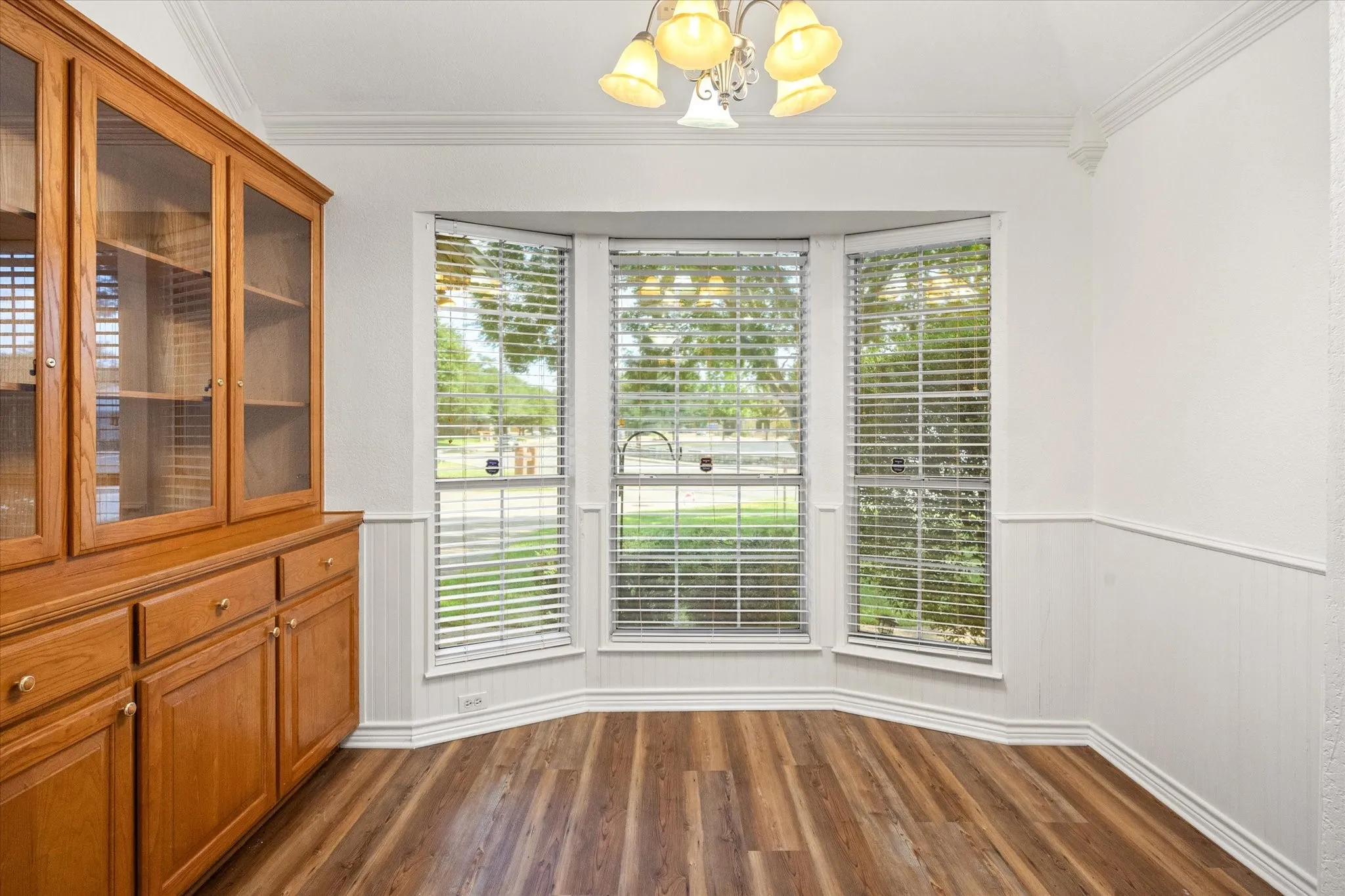 Bay window with blinds, wood-finish flooring, built-in cabinetry with glass display doors, decorative ceiling light fixture, and crown molding