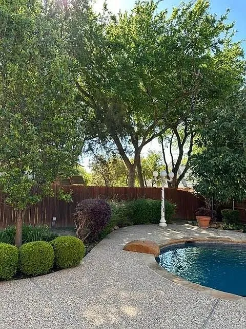 Swimming pool with a stone coping and exposed aggregate decking, surrounded by mature trees, privacy fencing, and manicured shrubs