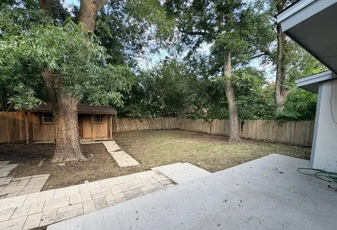 Expansive backyard featuring a concrete patio, mature trees, wood fence, and a wooden storage shed