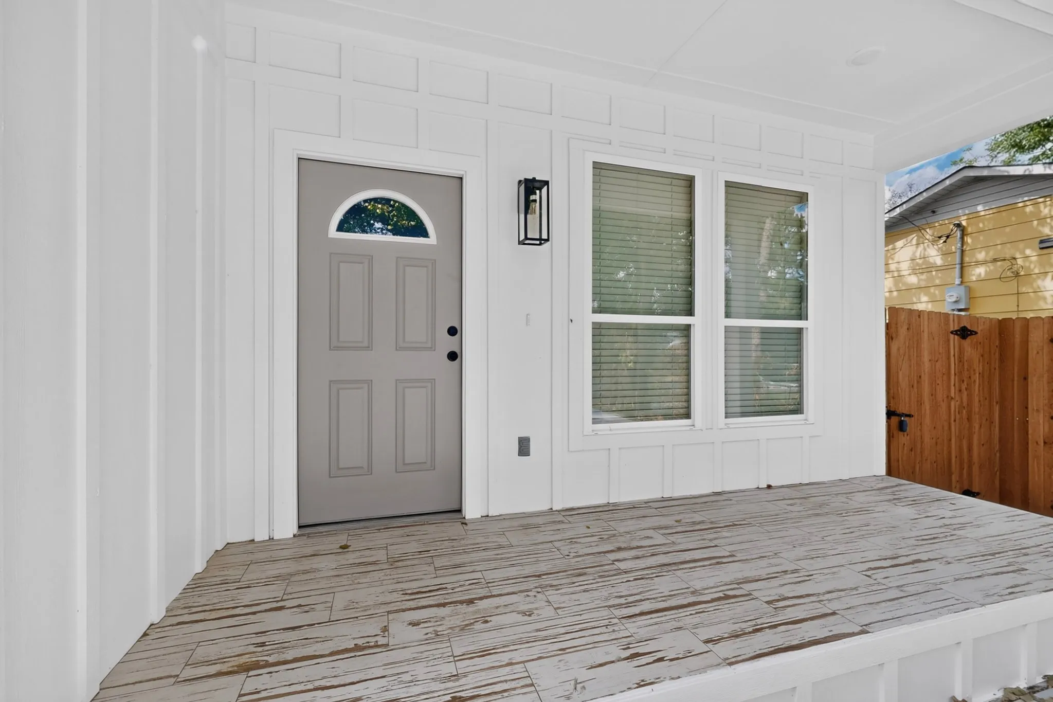 Front entry with a gray paneled door and half-moon glass insert, exterior wall lantern, and wood-finish porch flooring