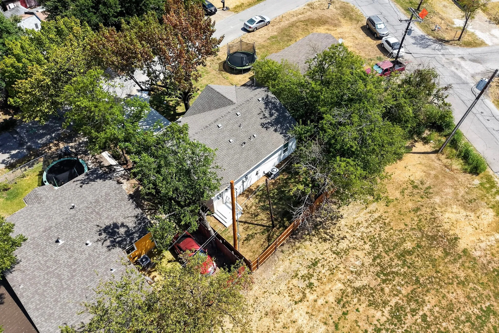 Aerial view of the property featuring a spacious asphalt driveway, mature shade trees throughout the grounds, and a detached structure with metal framing