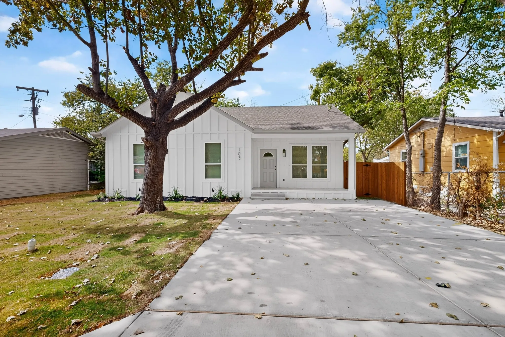 Modern white exterior with clean architectural lines, mature shade tree anchoring the front grounds, concrete driveway, attached garage, and manicured lawn with established landscaping
