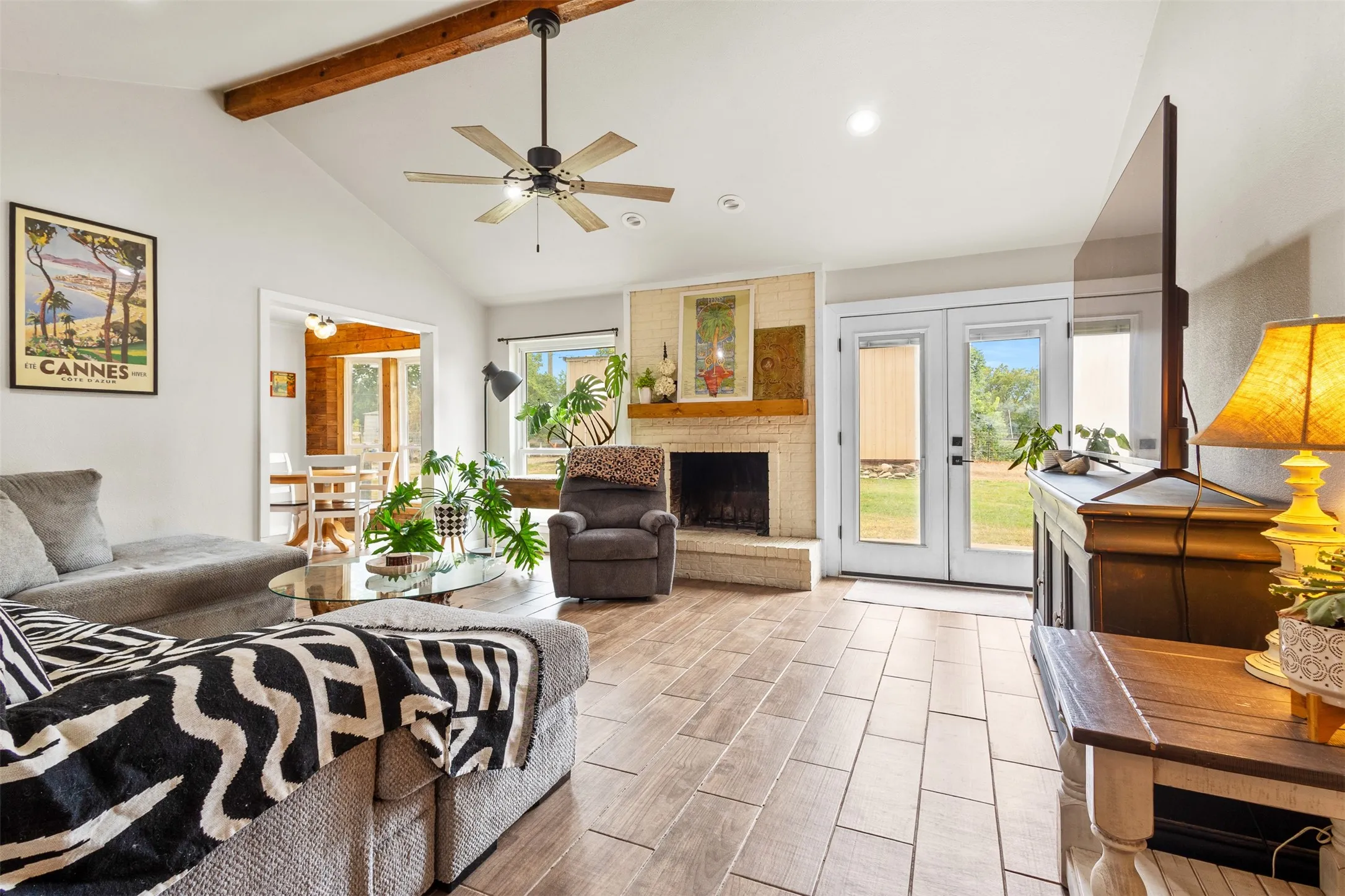 Vaulted ceiling with exposed wood beams and a ceiling fan, brick fireplace with a wood mantle, wood-finish tile flooring, and double glass-paneled doors