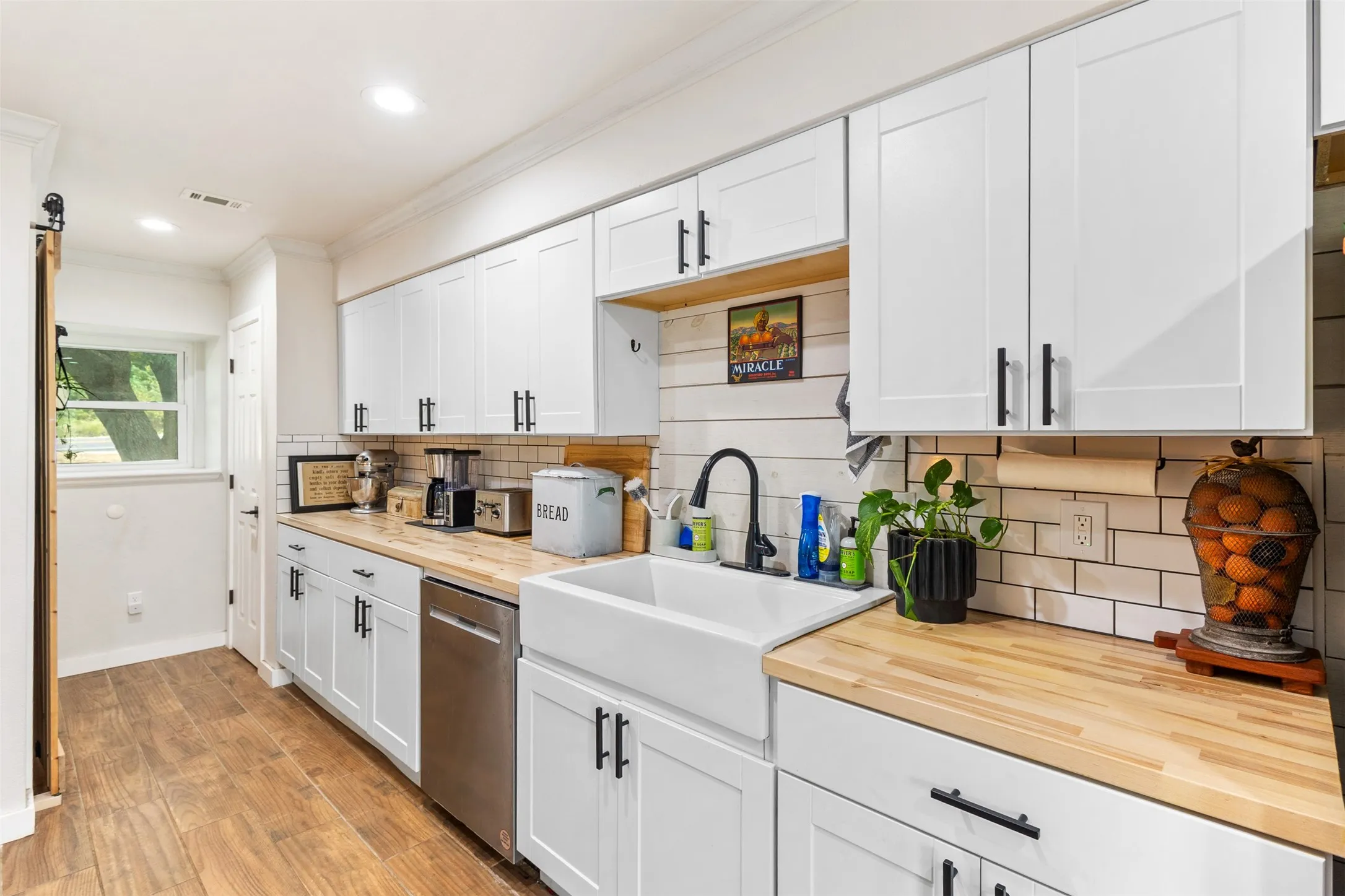 White shaker-style cabinetry with matte black hardware, wood-finish flooring, butcher block countertops, subway tile backsplash, and a farmhouse sink with a black gooseneck faucet