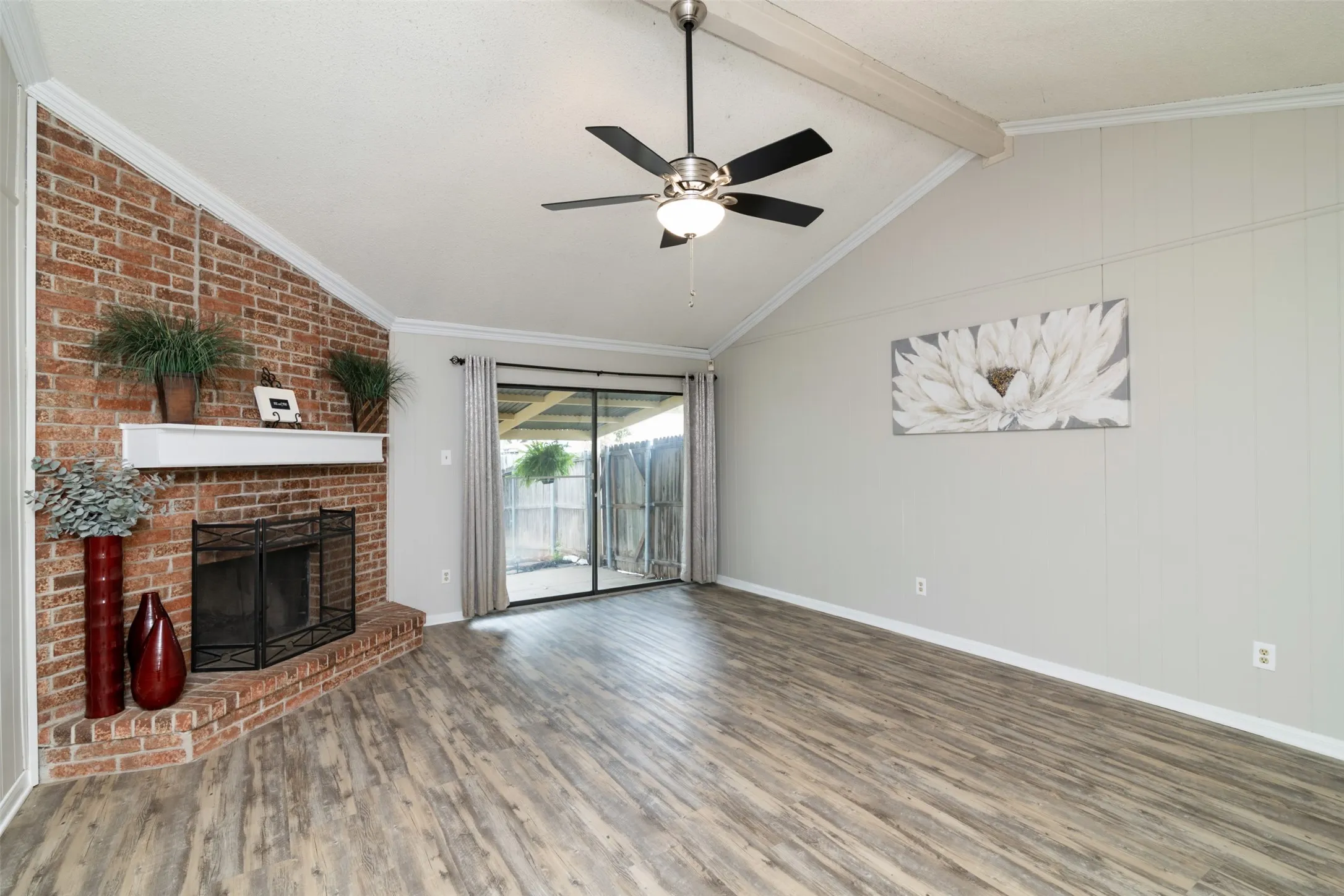 Vaulted ceiling with exposed beam, wood-finish flooring, brick fireplace with white mantel, ceiling fan with integrated lighting, sliding glass​​‌​​​​‌​​‌‌​‌‌​​​‌‌​‌​‌​‌​​​‌​​ door