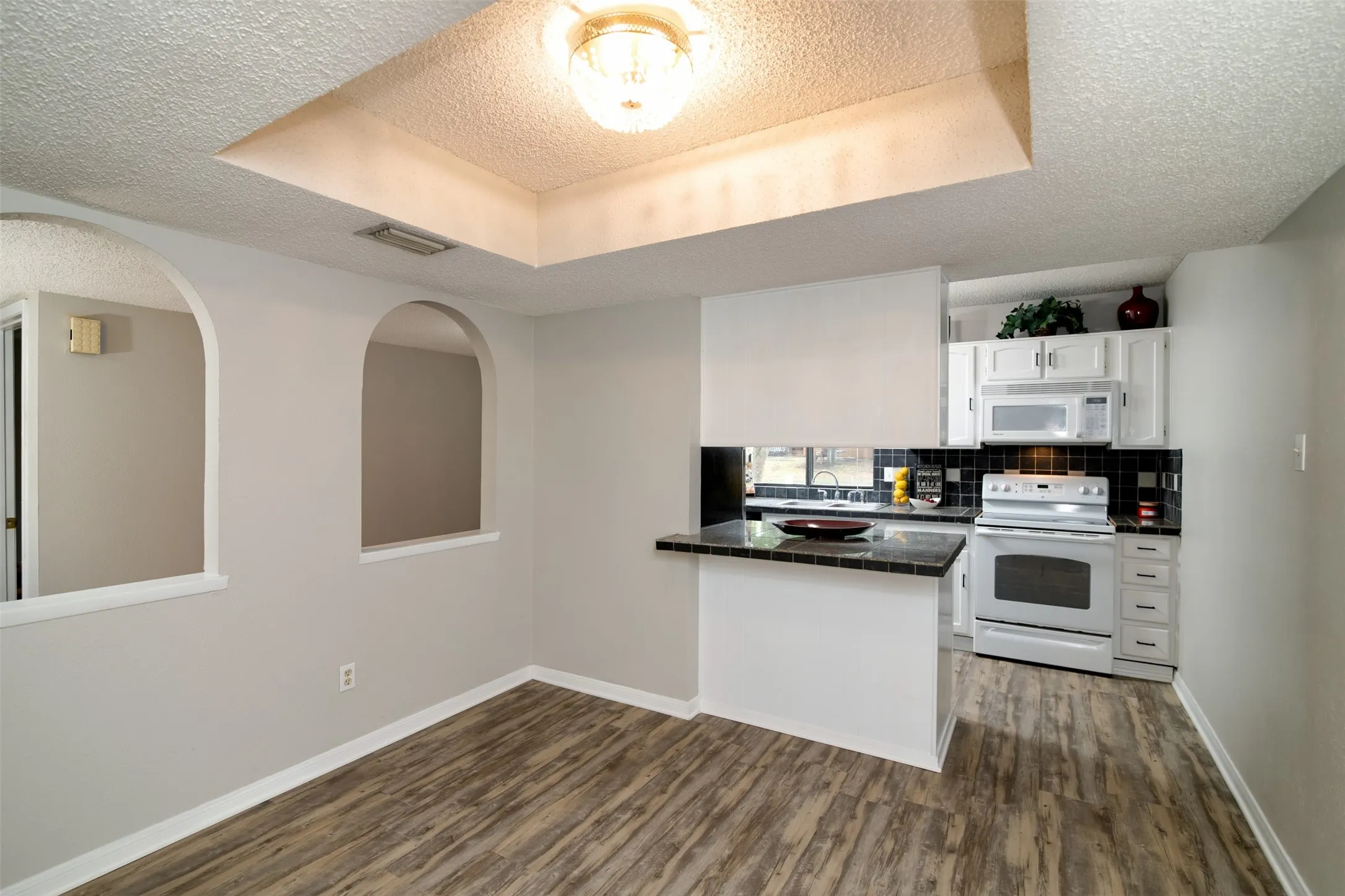 Recessed ceiling with an overhead light fixture, wood-finish flooring, arched wall openings, a kitchen pass-through with a breakfast bar, and white cabinetry