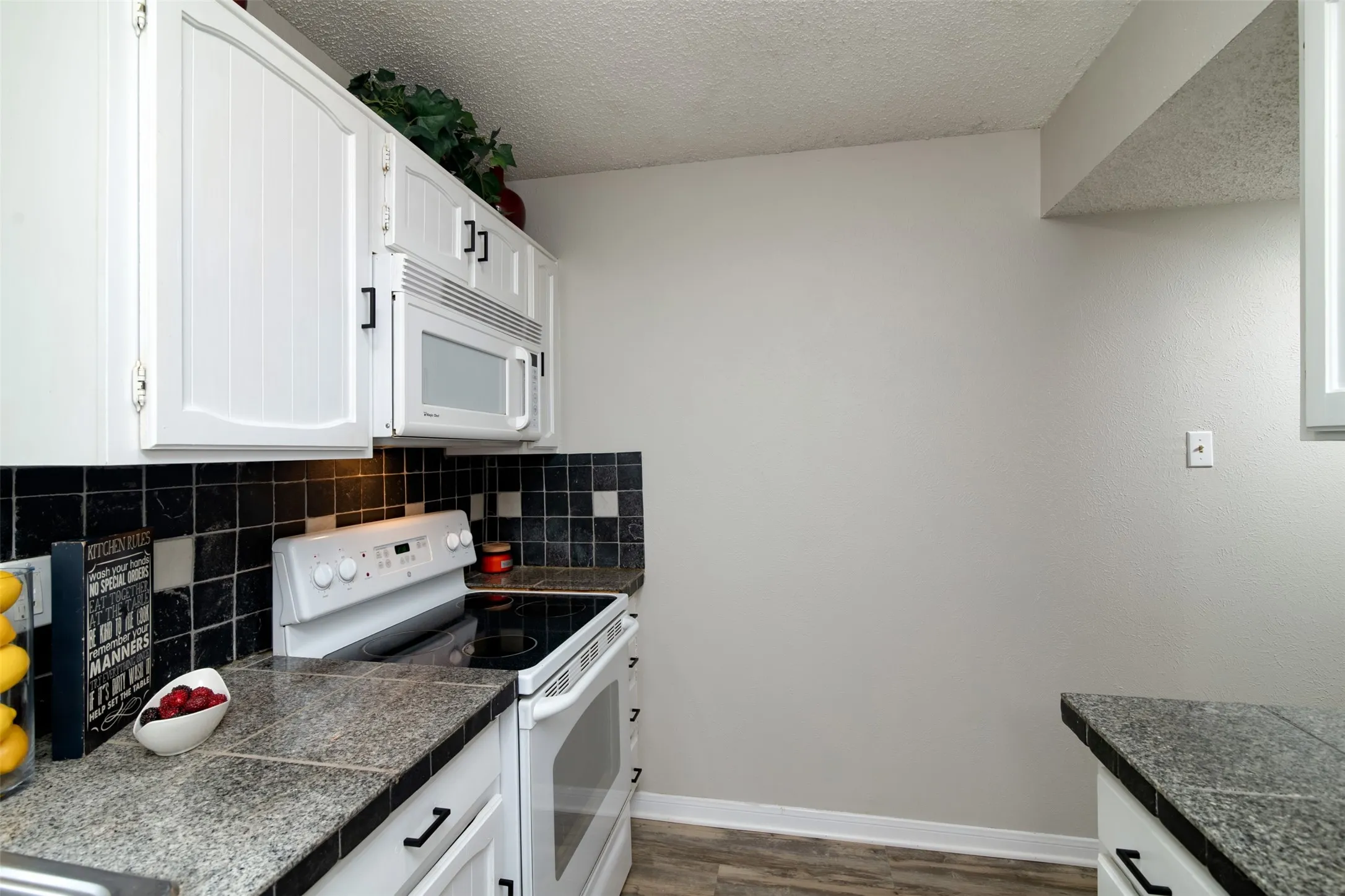 White cabinetry with black hardware, stone-finish countertops, black and white tile backsplash, and wood-finish flooring