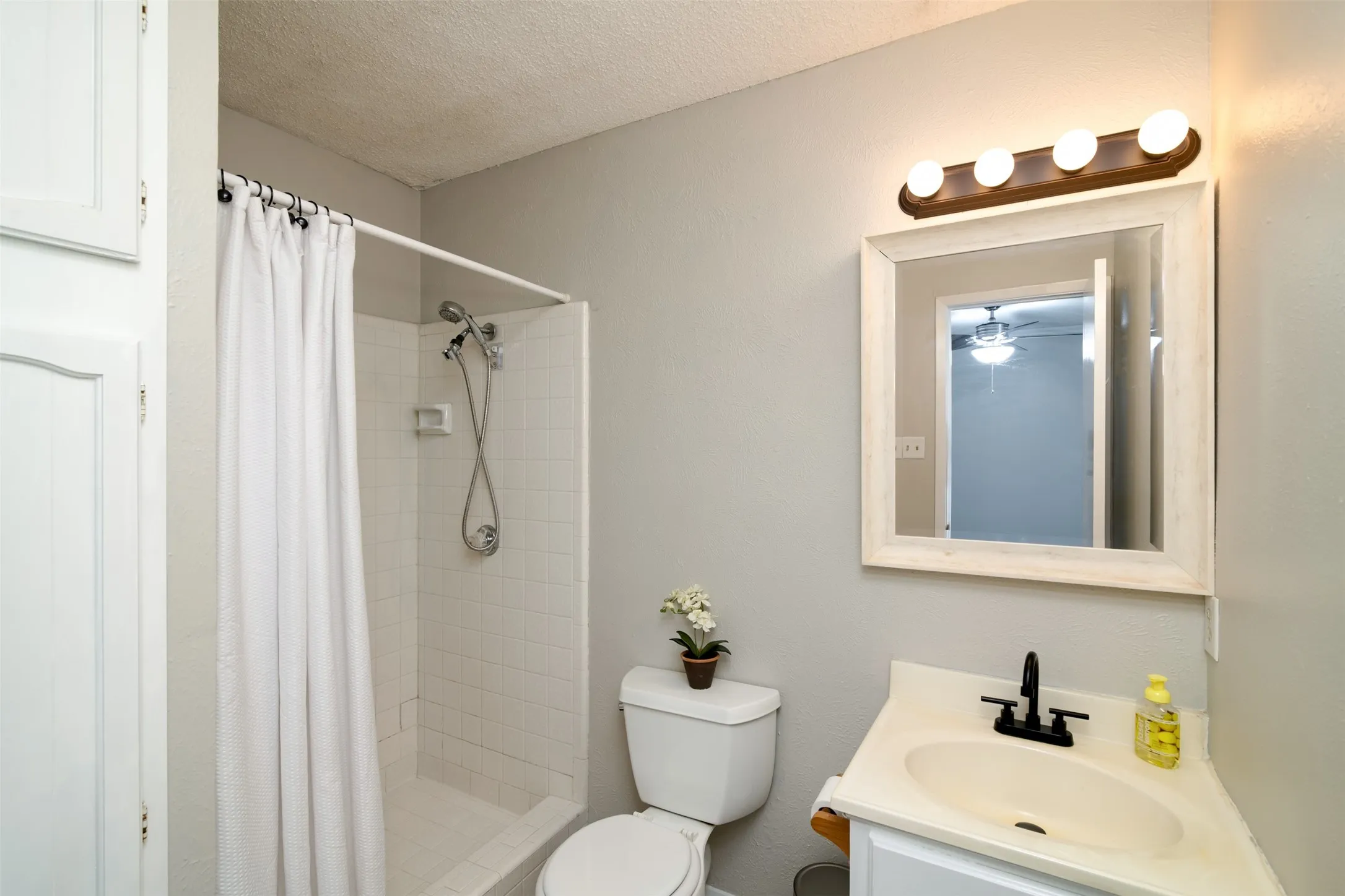 Shower with white tiled surround, corner vanity with undermount sink, white framed mirror, and a four-bulb vanity light fixture